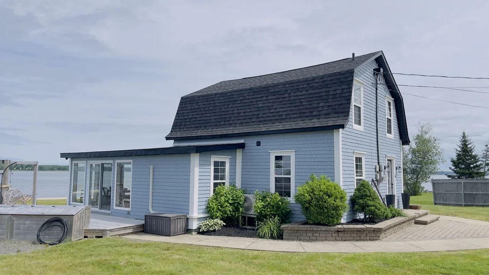 Side view of a repainted light blue gambrel cottage with white window trim near the water in Shediac