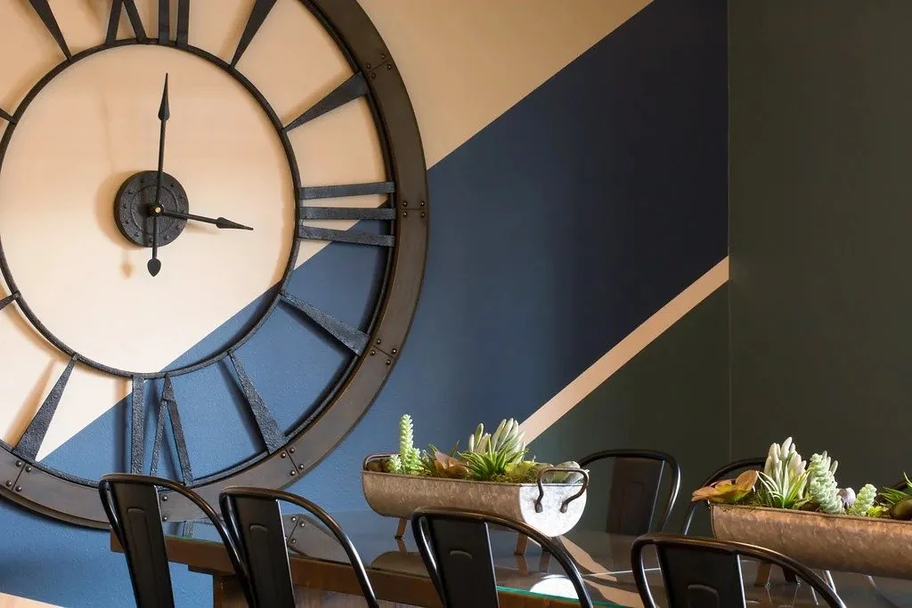 A large analog clock hung on a blue, green, and white striped wall in a dining room.