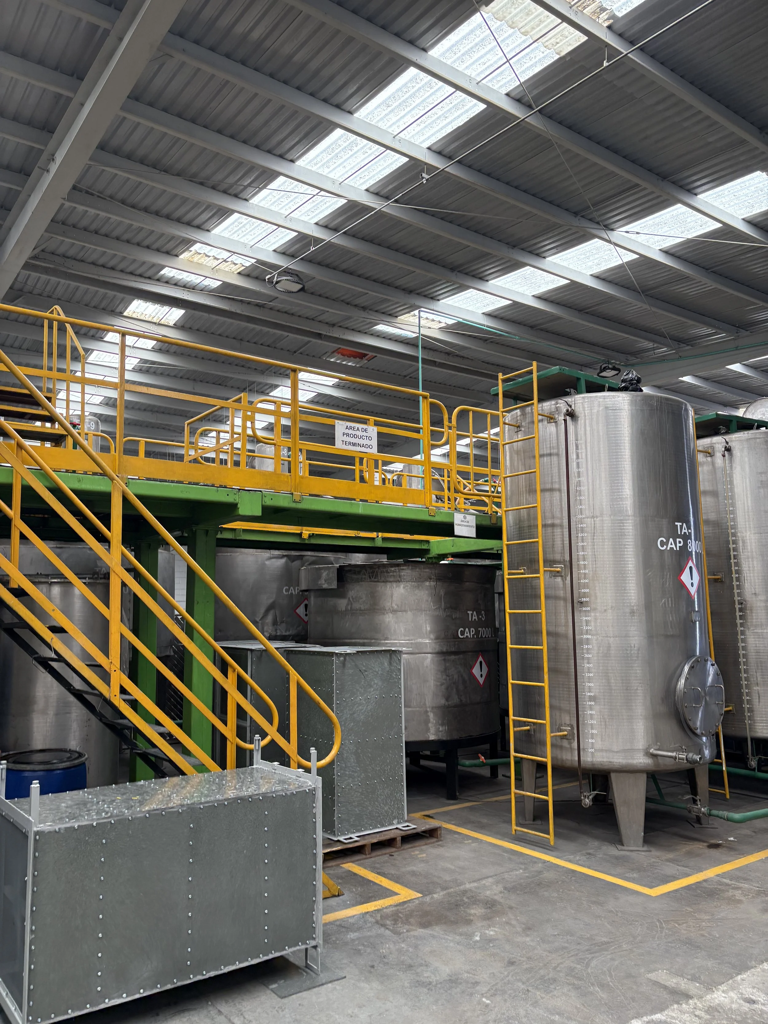 Industrial facility with large stainless steel tanks, yellow safety railings, and metal staircase, used for processing or storing liquids.