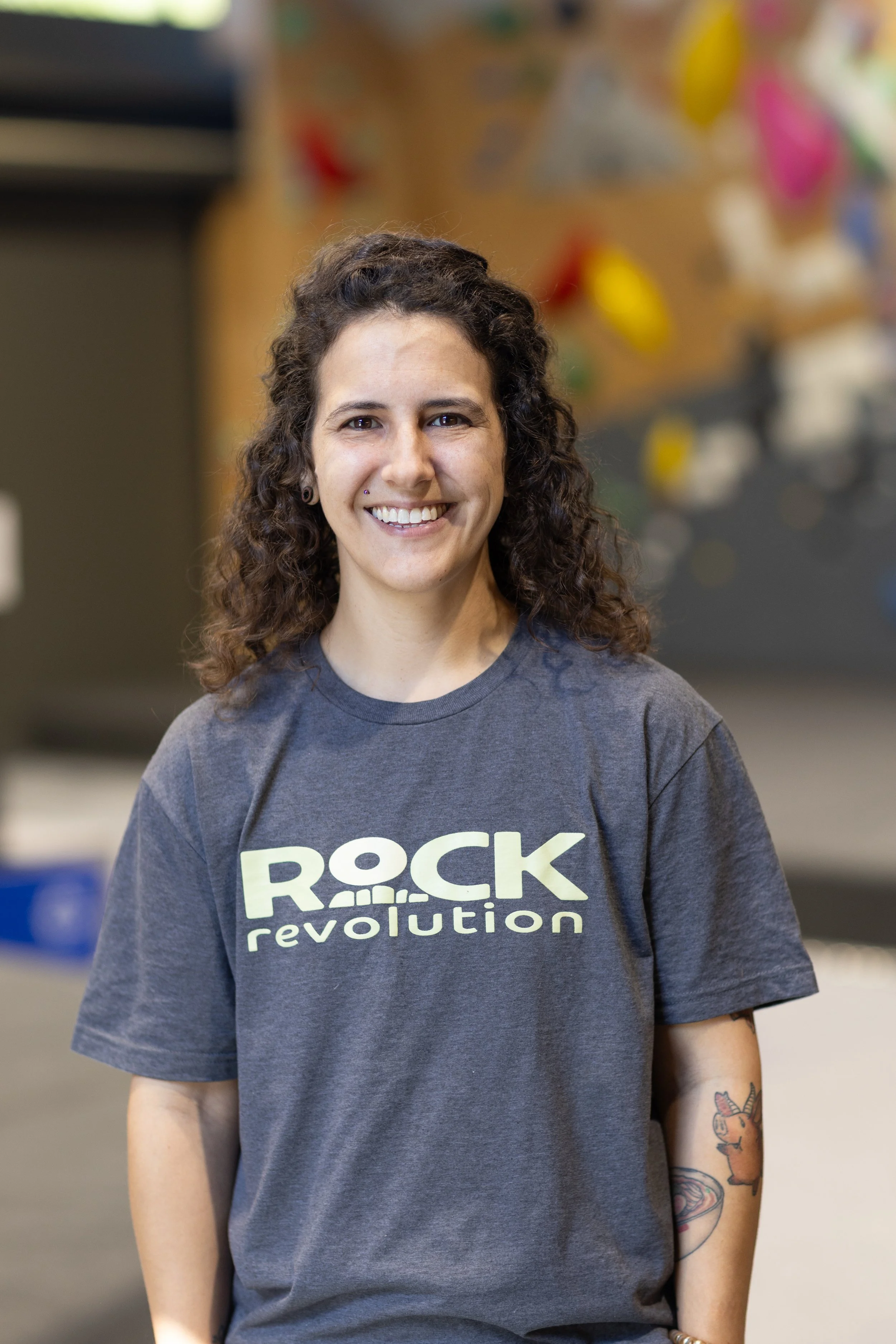 A woman with curly brown hair, smiling, wearing a gray T-shirt with the words 'Rock Revolution' printed on it, standing indoors in a rock climbing gym.