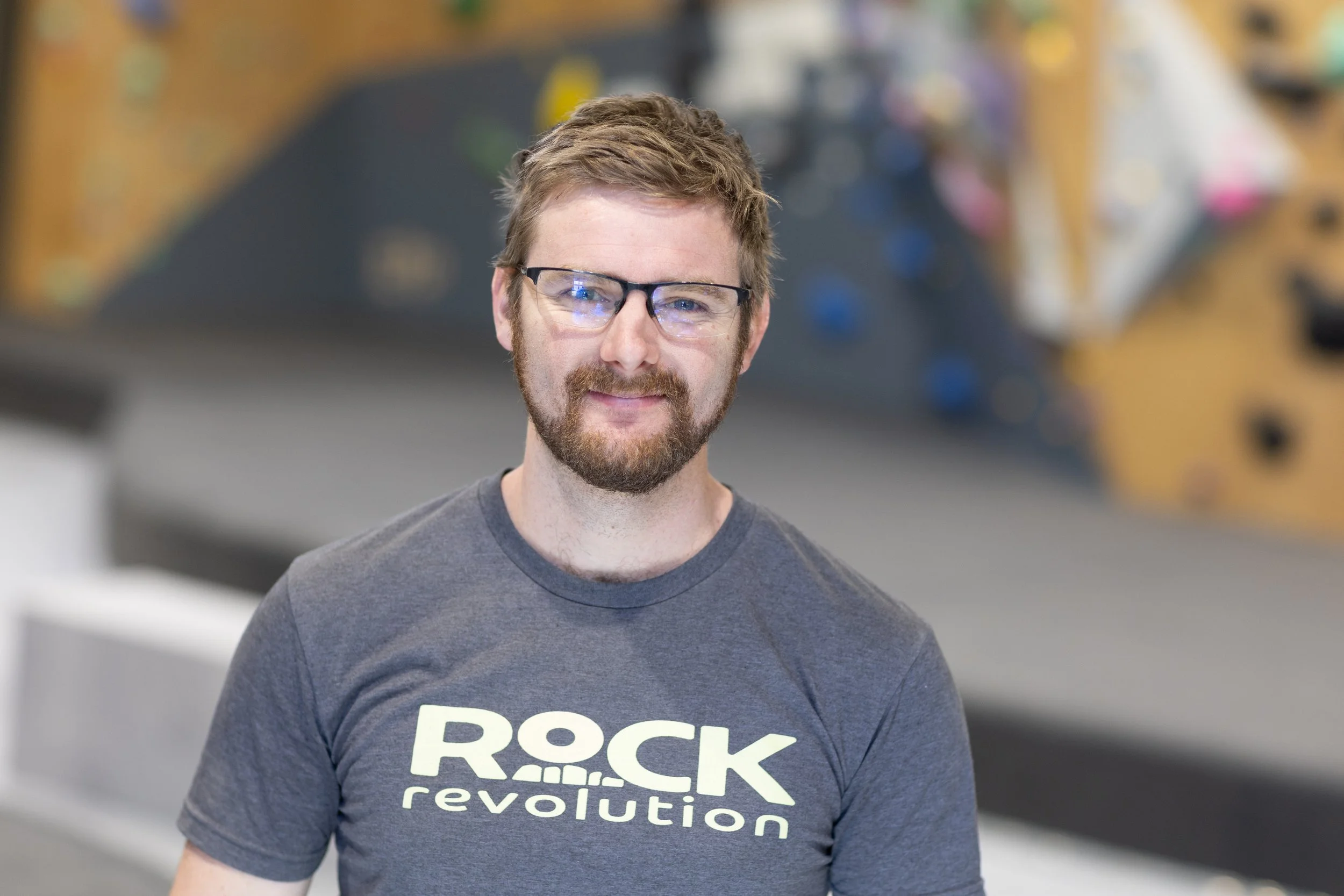 Man with glasses and beard smiling, wearing a dark gray T-shirt that reads 'Rock Revolution', in an indoor rock climbing gym.