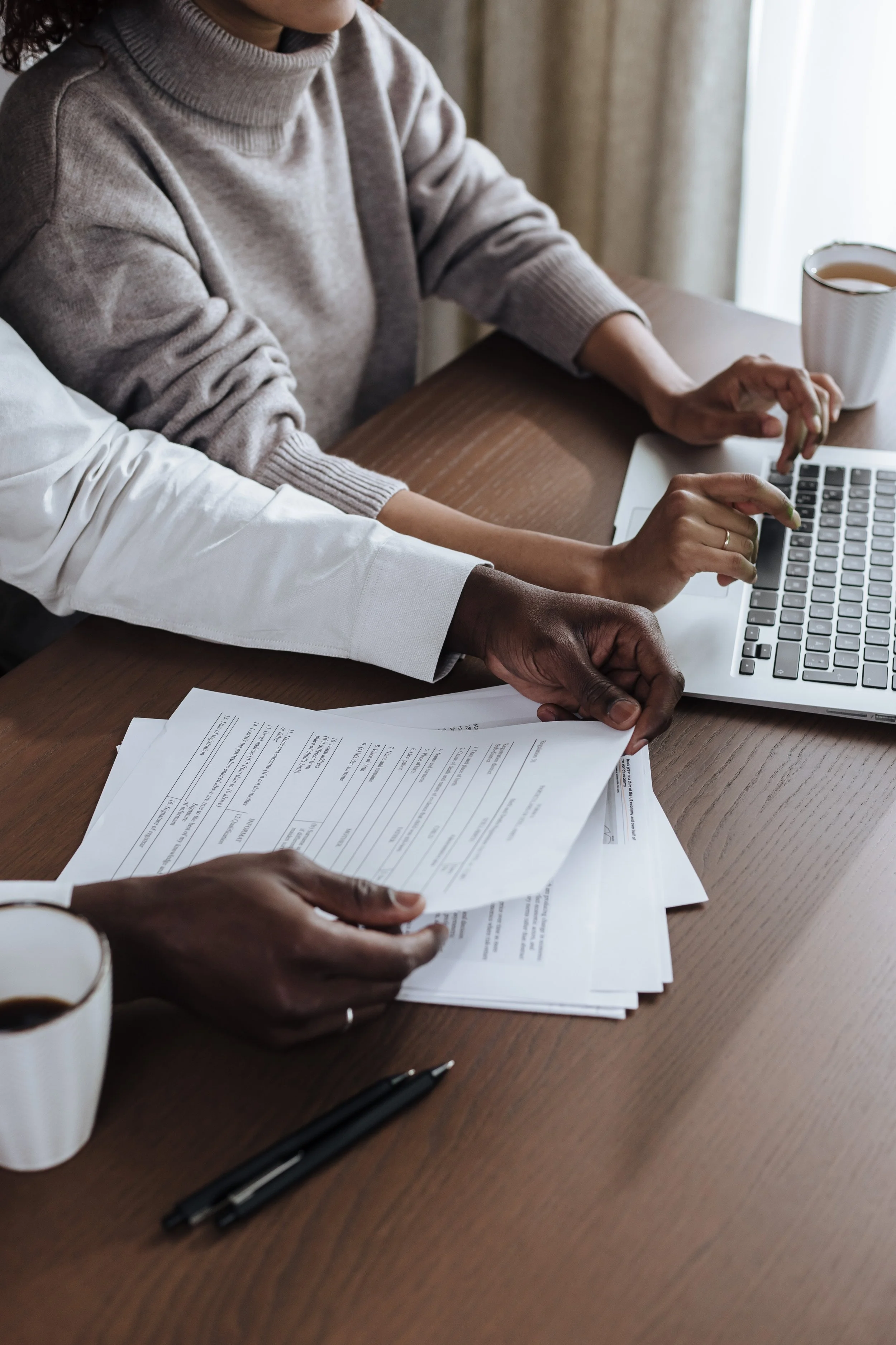 Two people working together at a desk with paperwork, a coffee cup, a laptop, and a pen.