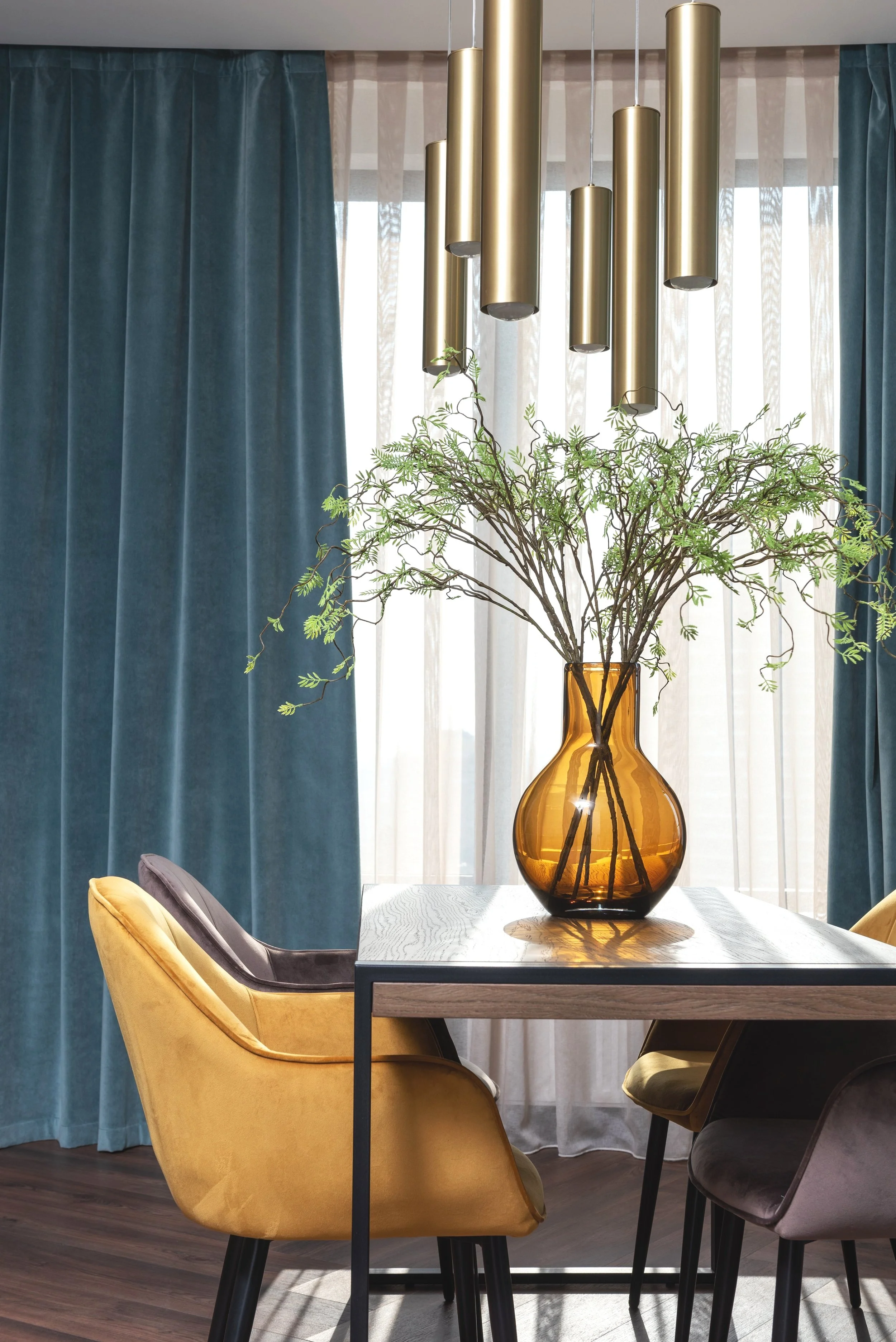 A dining room with a wooden table, yellow and gray chairs, a glass vase with green branches, teal curtains, sheer curtains, and hanging gold pendant lights.