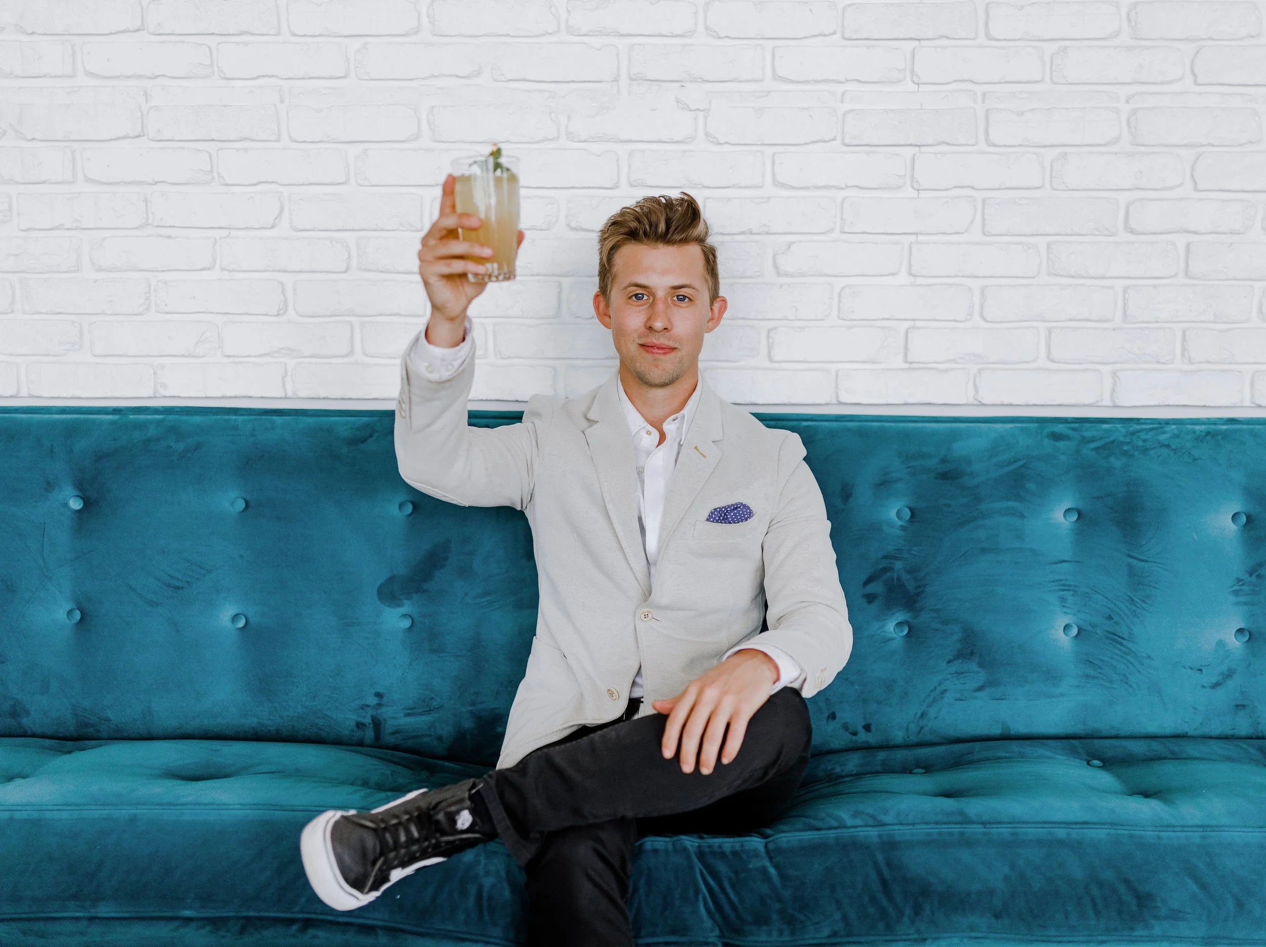 A young man in a light gray blazer and white shirt sitting cross-legged on a teal velvet couch, holding up a cocktail glass with a drink and garnish, against a white brick wall.