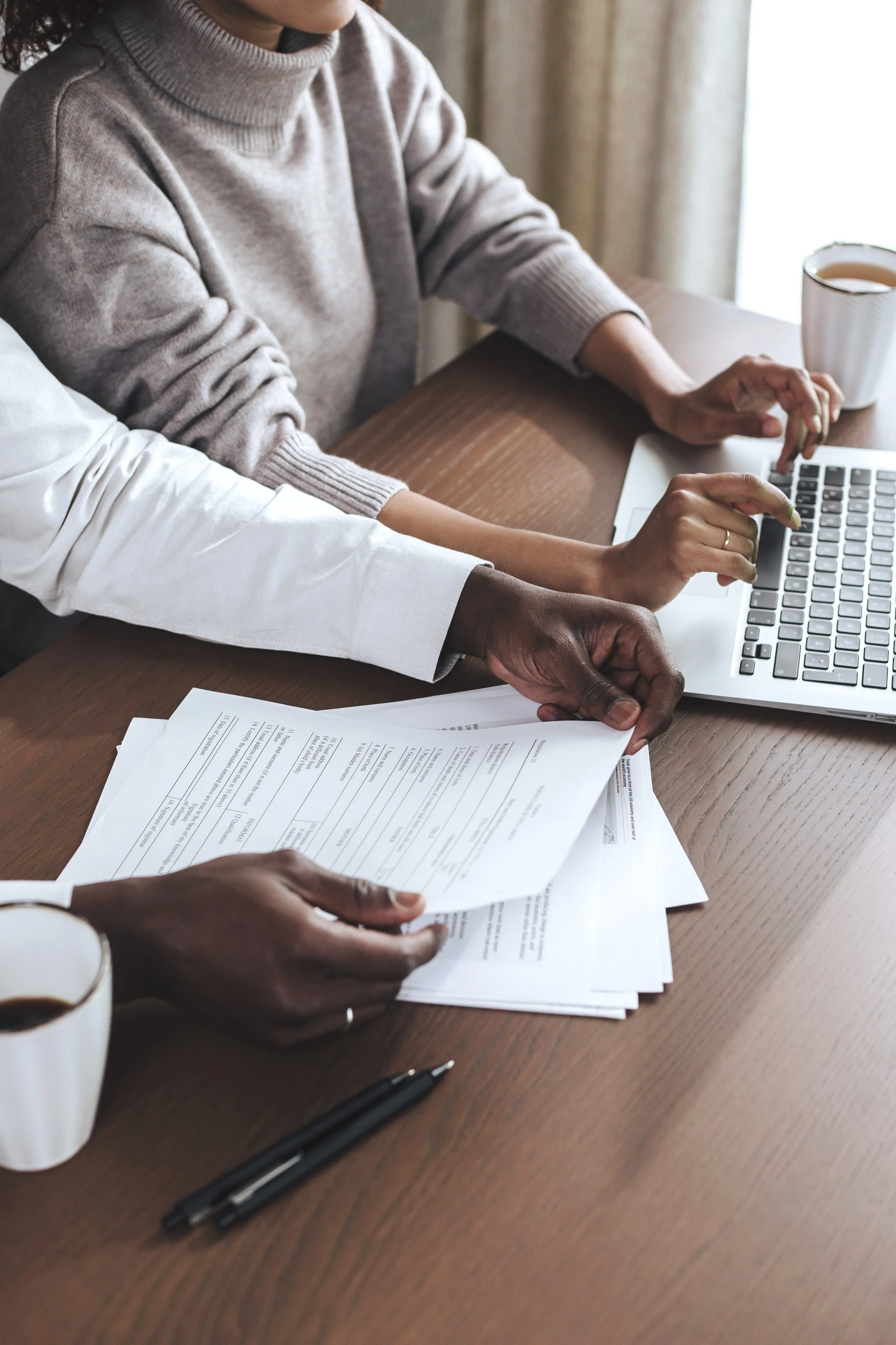 Two people working together at a wooden table, reviewing documents and using a laptop, with coffee cups nearby.