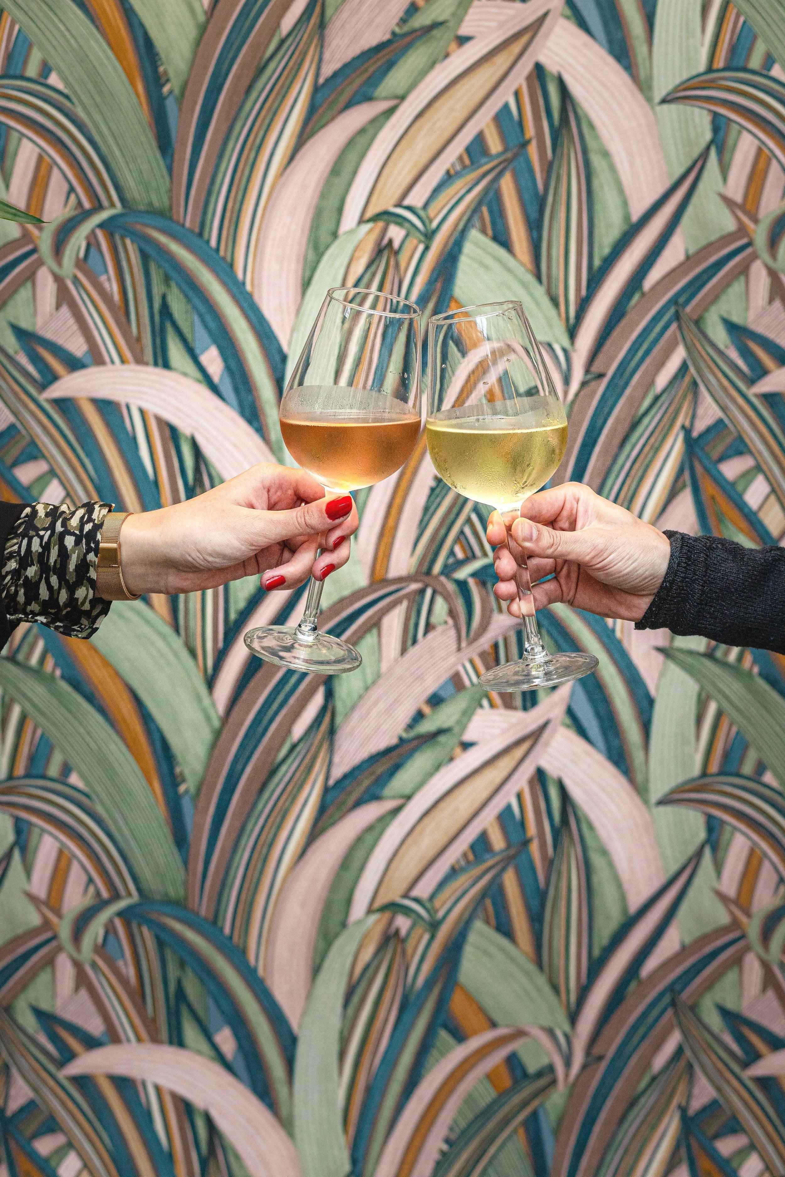 Two people clinking glasses of white wine against a colorful leaf-patterned wall.