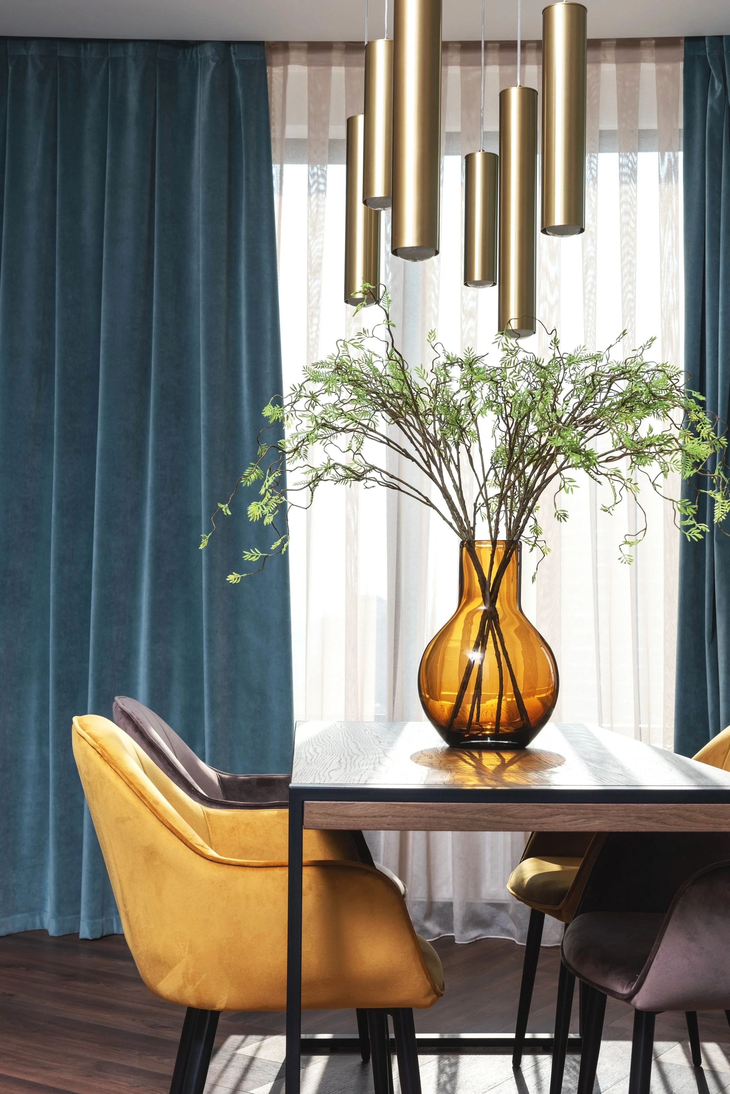 A dining room with teal curtains, a wooden table, and a large amber glass vase holding green leafy branches, with modern gold pendant lights overhead.