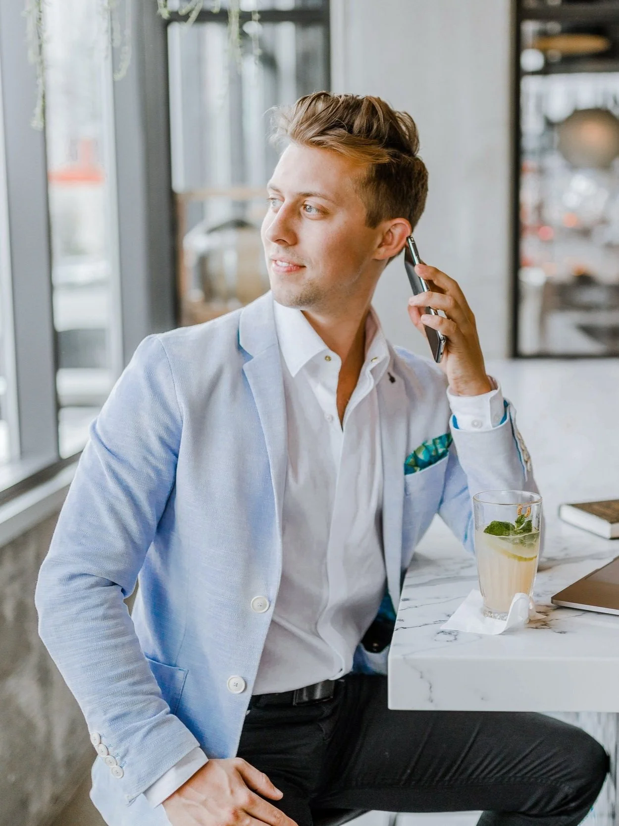 A young man in a light blue blazer and white shirt sits at a white marble table in a cafe, holding a phone to his ear, with a drink and laptop on the table.