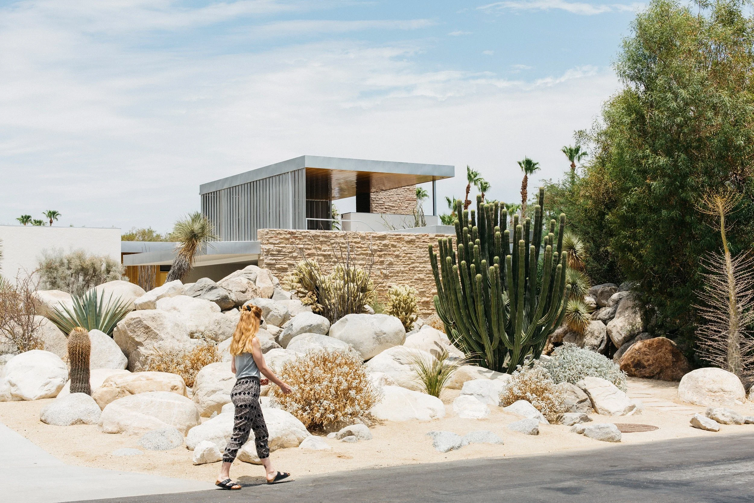 A woman with blond hair walking in a desert landscape with large rocks, cacti, and desert plants, in front of a modern house with a flat roof and metallic siding.