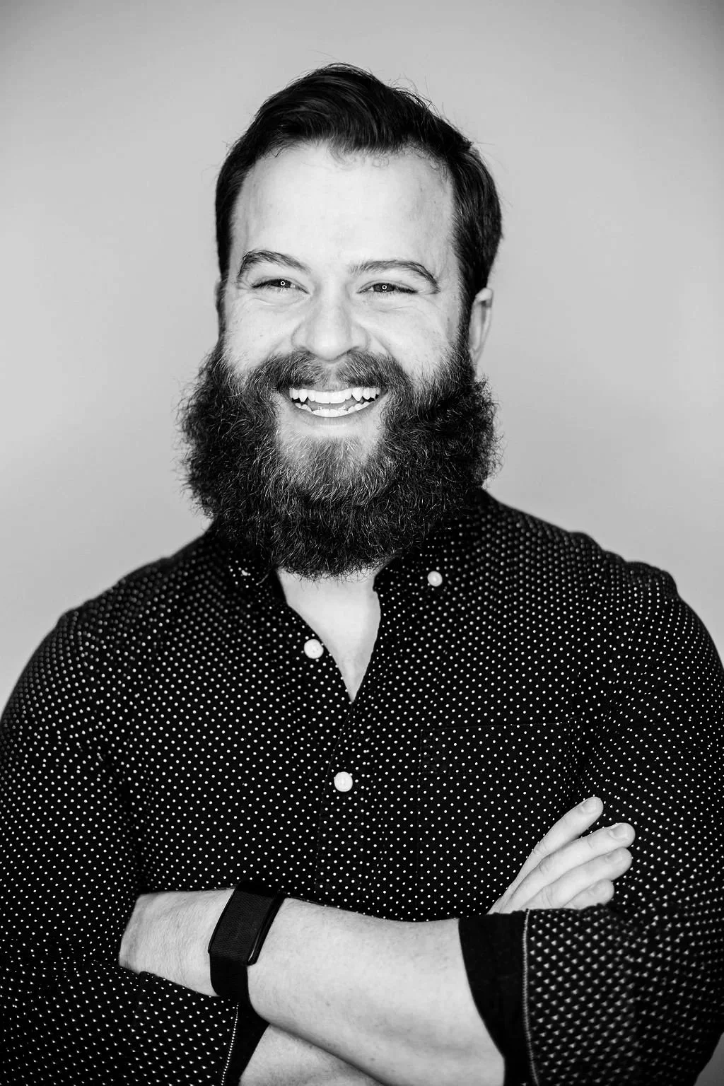 A smiling man with a beard and mustache, wearing a dark polka-dotted shirt, with arms crossed, in front of a plain background.