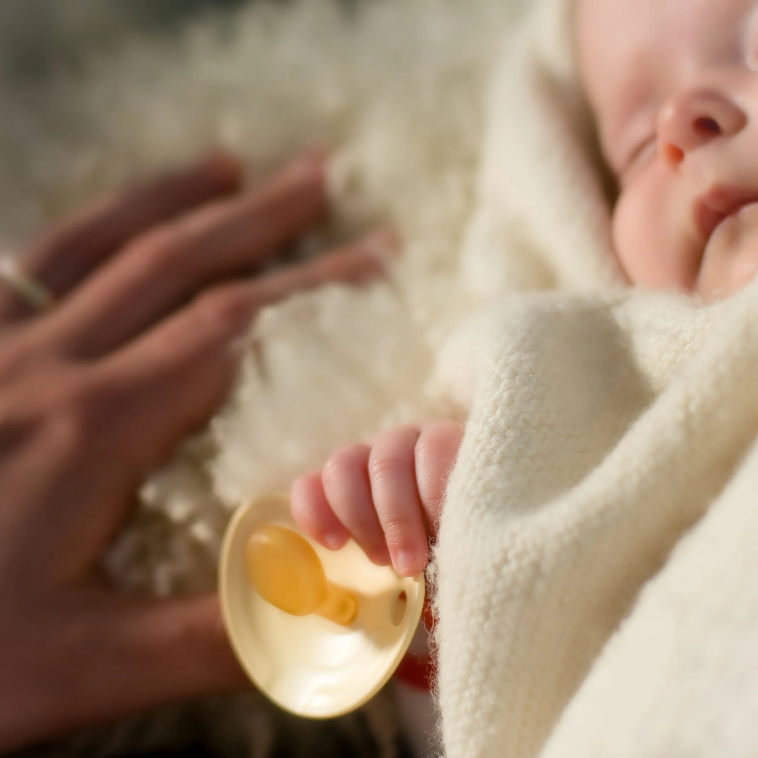 Nahaufnahme eines neugeborenen Babys, das in einer weichen, cremefarbenen Decke liegt, während eine Hand einen Schnuller hält.