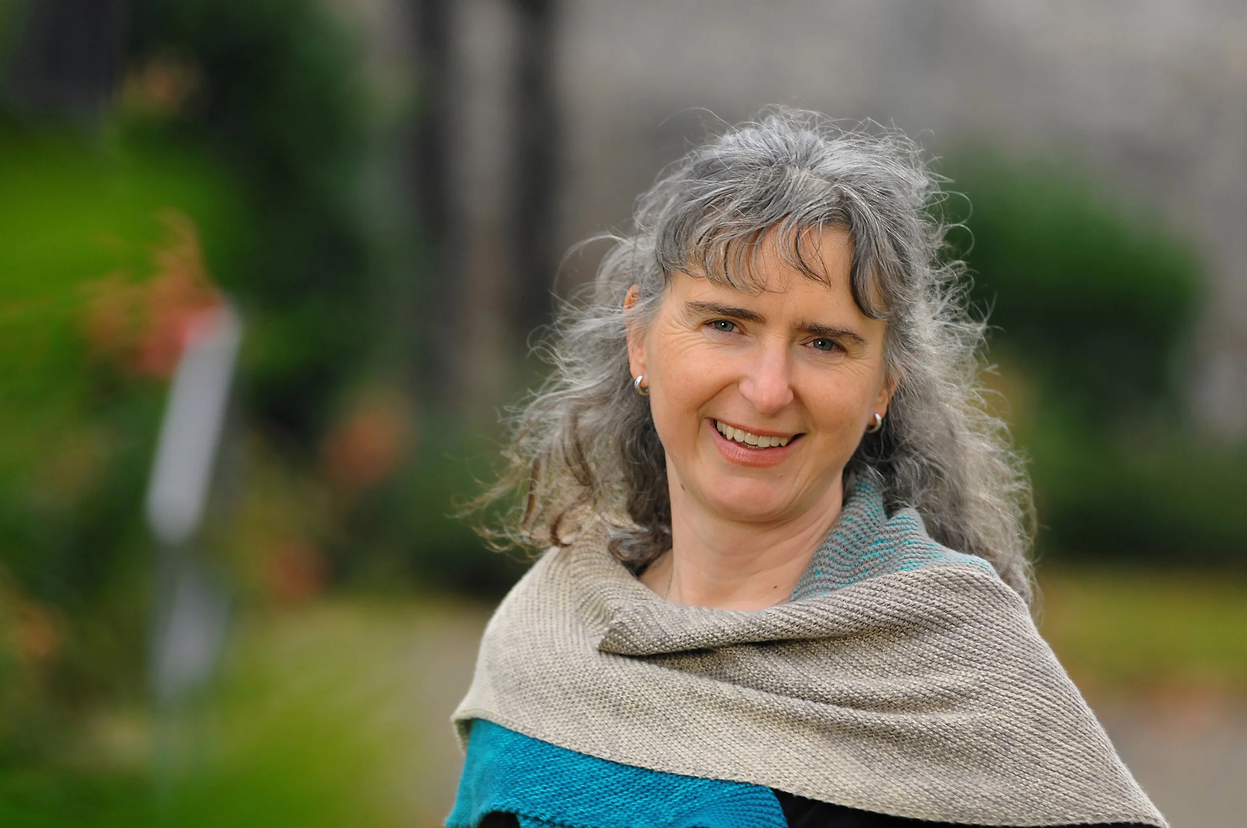 Eine lächelnde Frau mit silbergrauem, lockigem Haar, trägt einen beige-grauen Schal und eine blaue Jacke, steht im Garten mit grünem Unkraut im Hintergrund.