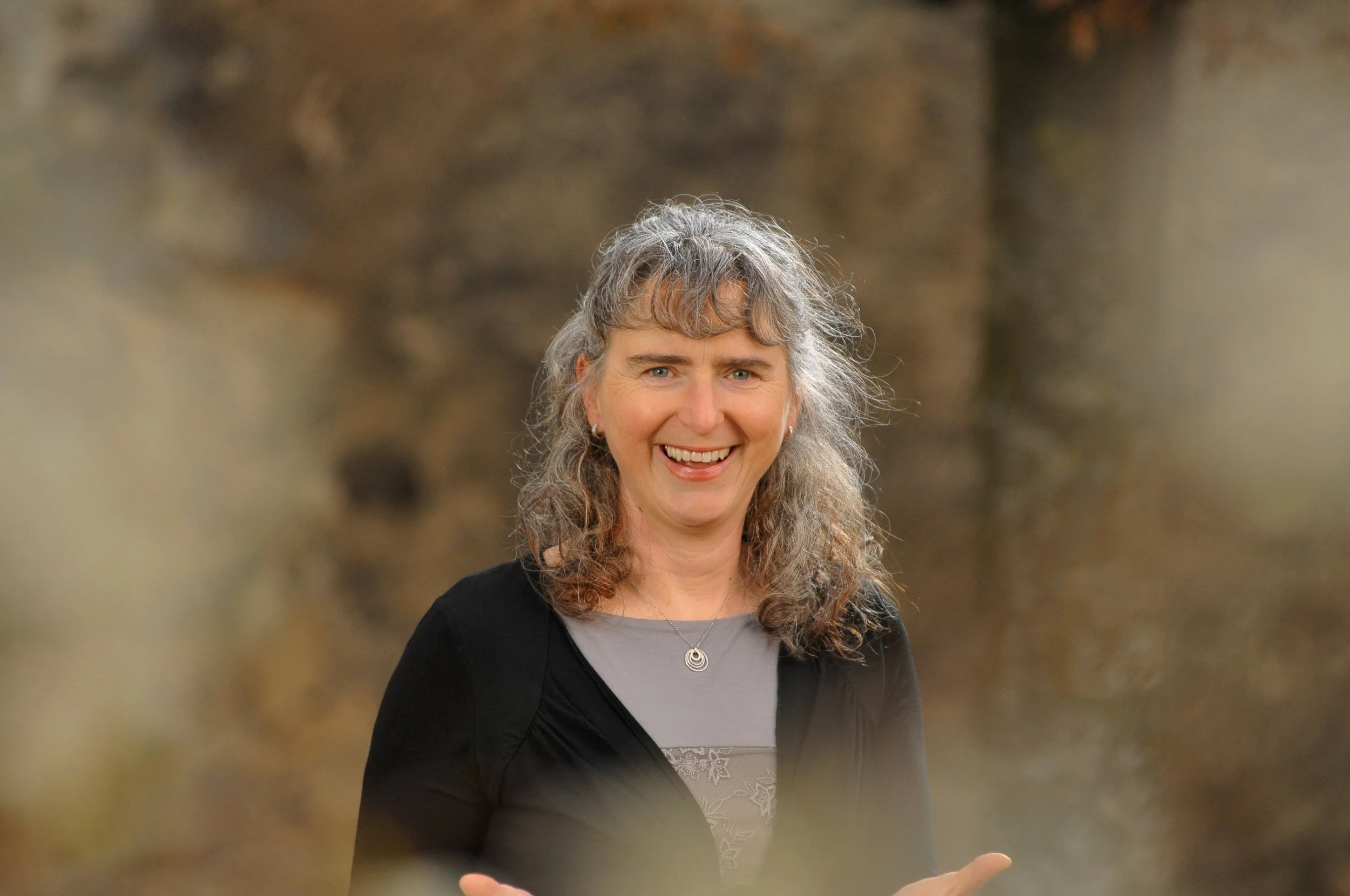 Eine lachende Frau mit grauem, leicht gewelltem Haar, Trägerin eines schwarzen Jäckchens und einer hellgrauen Bluse, vor einer unscharfen natürlichen Hintergrundkulisse, hält Arme im 90° Winkel mit Handflächen nach oben