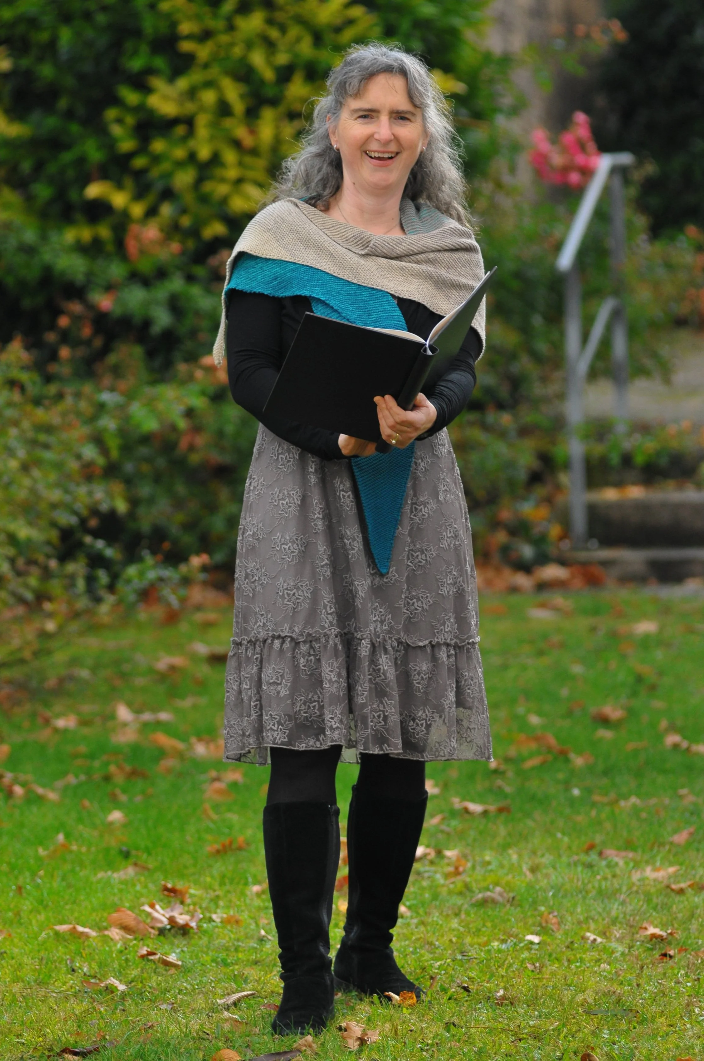 Freie Rednerinn (IHK): Eine lachende Frau mit grauem Haar, die eine Rednermappe hält, steht auf einem grünen Rasen im Freien, umgeben von Herbstlaub und Büschen.
