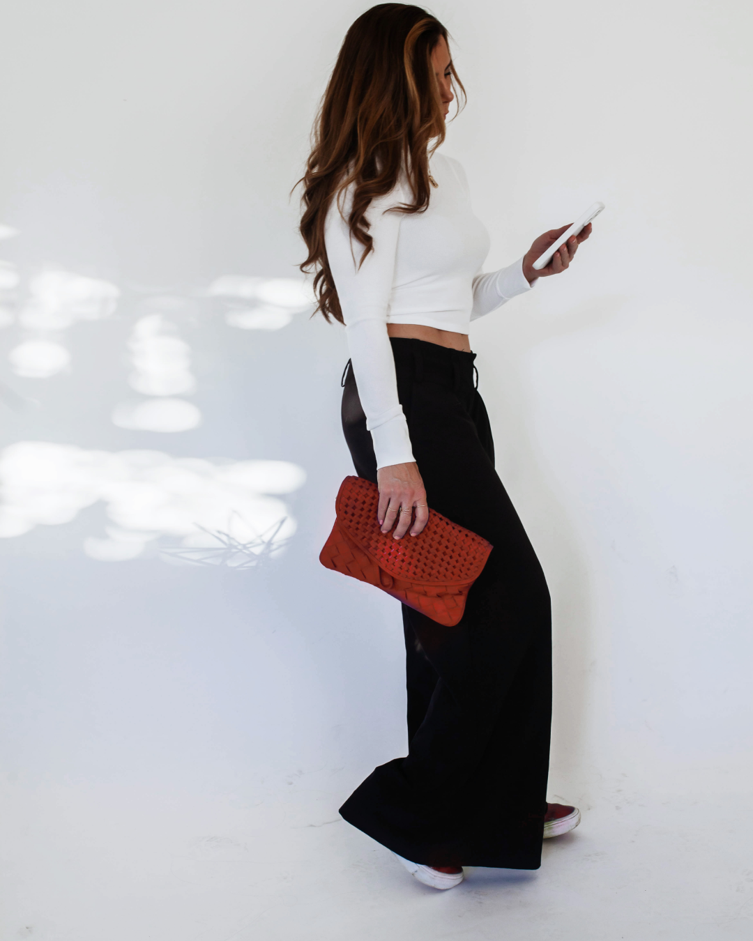 Woman with long, wavy brown hair, standing against a white wall, looking at her phone, dressed in a white long-sleeve crop top, black wide-leg pants, holding a red woven clutch.