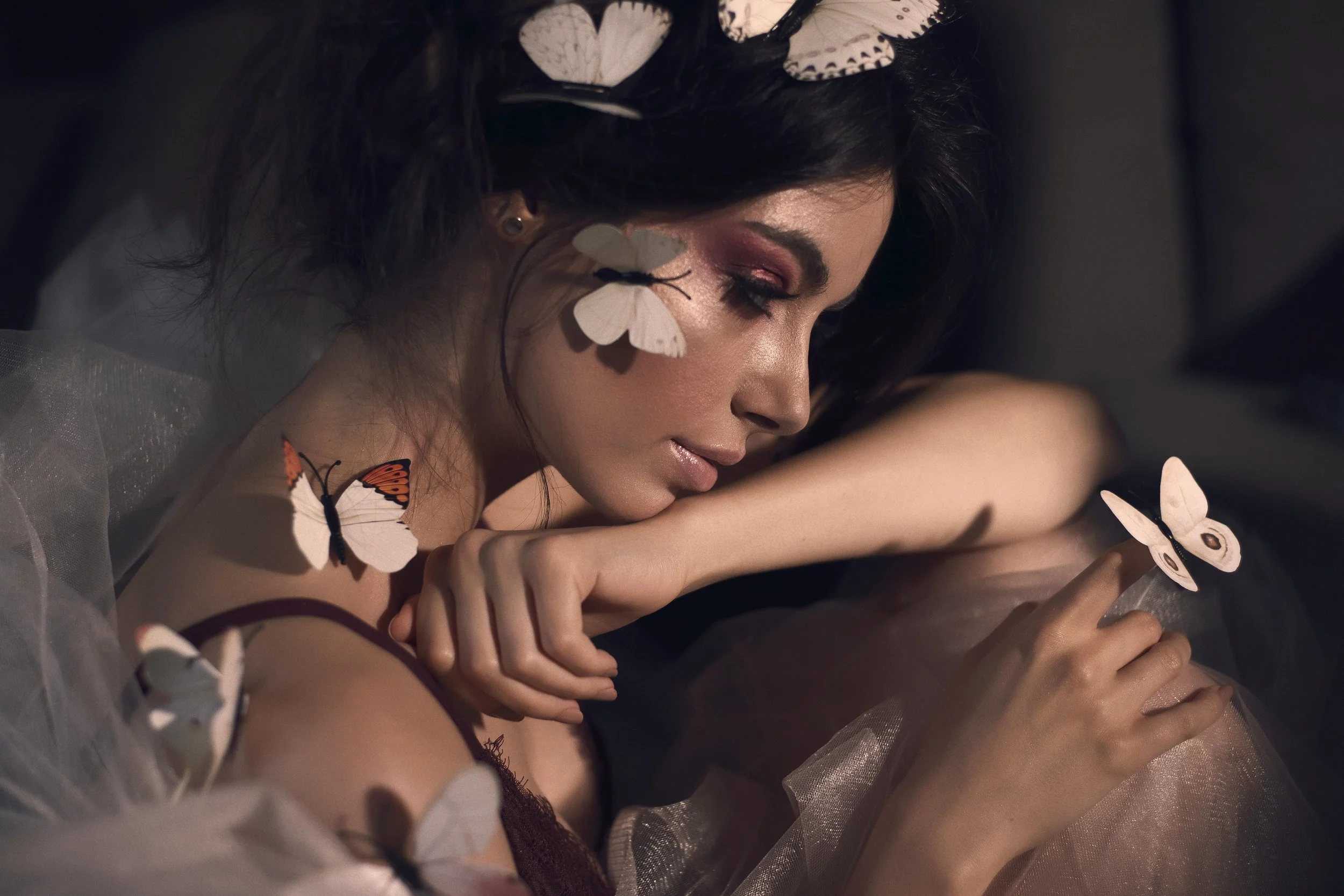 A woman with dark hair adorned with white butterfly hair accessories, sitting with her eyes closed, surrounded by white butterfly cutouts on her shoulder, arm, and nearby. She has pink eyeshadow and a serene expression.