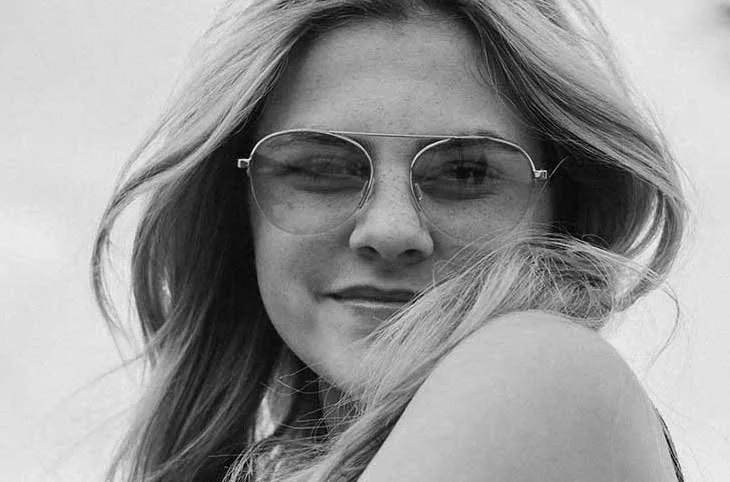 A young woman with long hair wearing sunglasses, smiling and winking at the camera, in black and white.