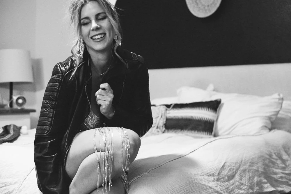 A woman with curly hair smiling and laughing while sitting on a bed, wearing a leather jacket and holding a necklace with long beads.