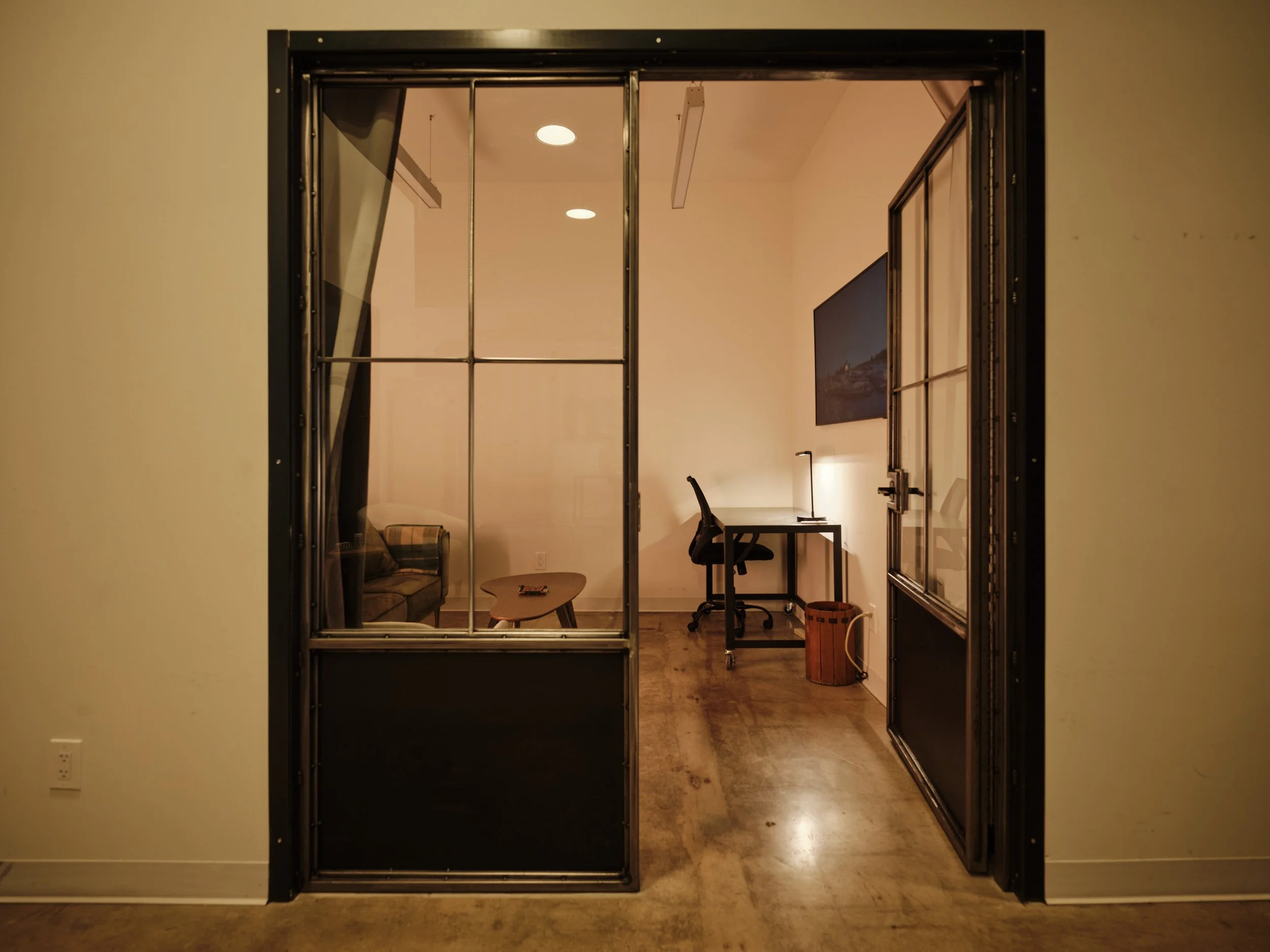 View of a small room with a glass-pane door, featuring a black frame, leading into a minimalist office space with a desk, chair, lamp, and a wall painting. Through the door, part of a living area with a sofa and coffee table is visible.