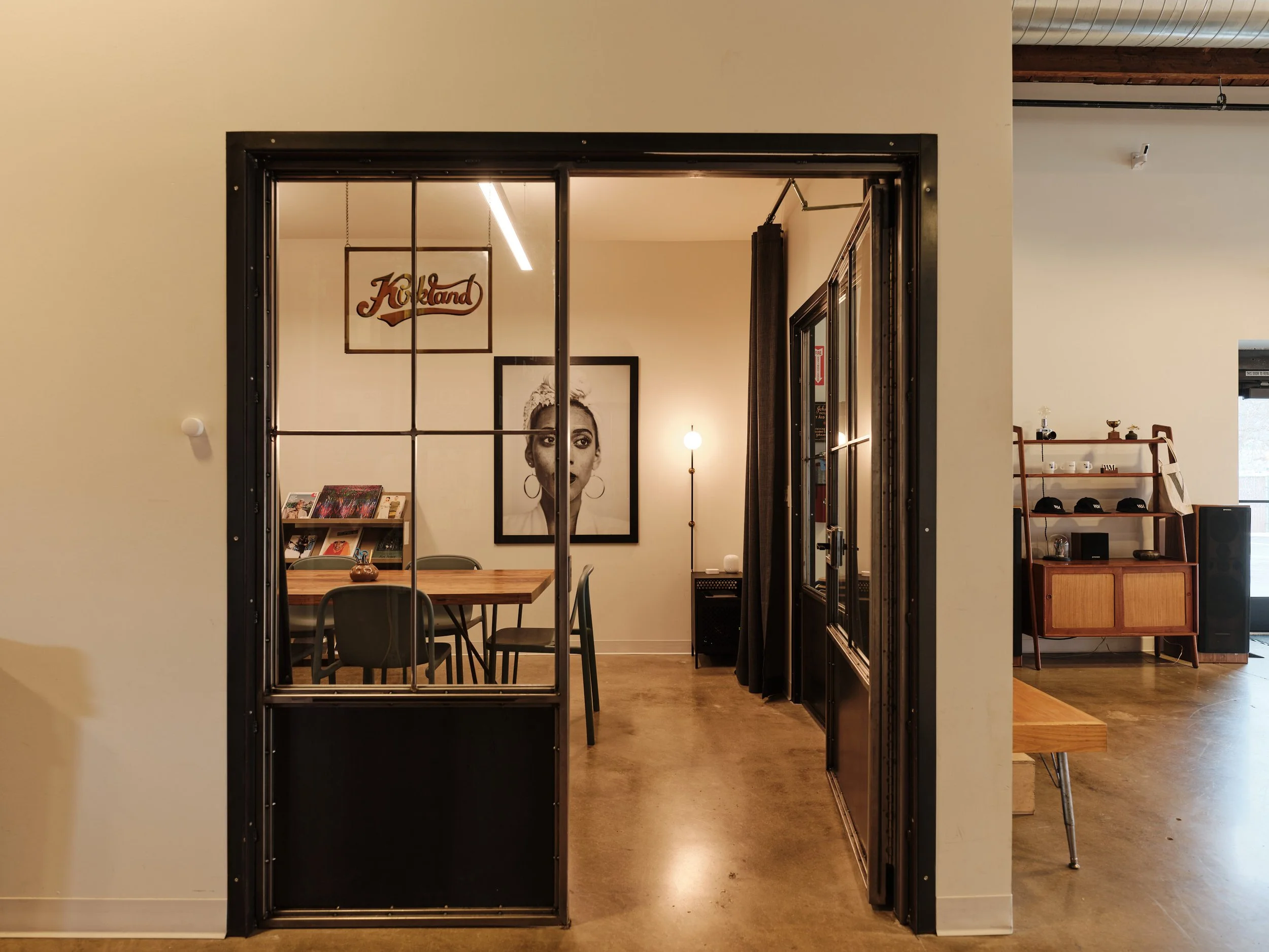 View into a meeting room through a metal-framed glass door, featuring a wooden table, chairs, artwork on the wall, and a floor lamp.