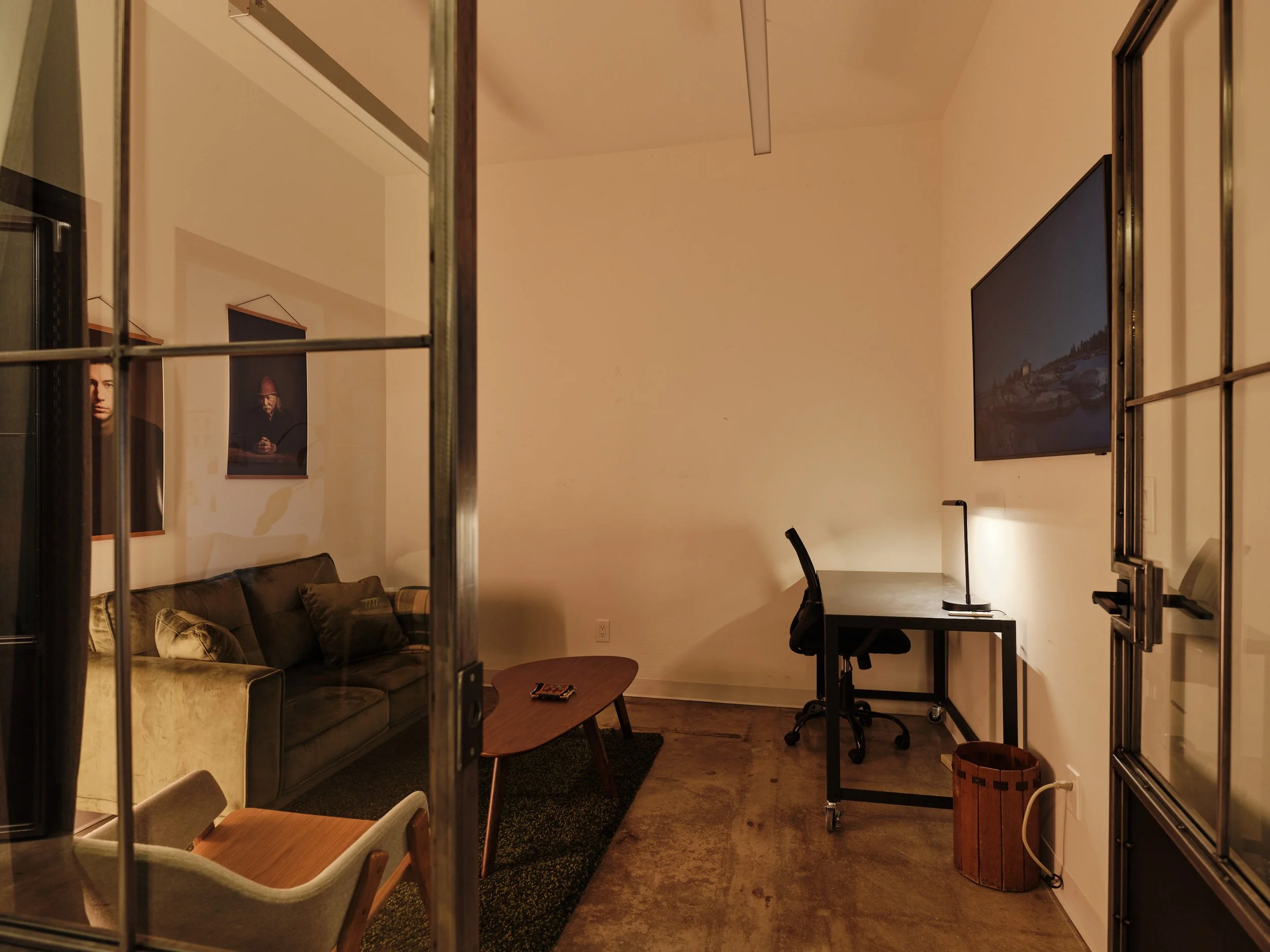 Living room with a sofa, armchair, coffee table, desk, and wall-mounted TV, viewed through a glass-paned door.