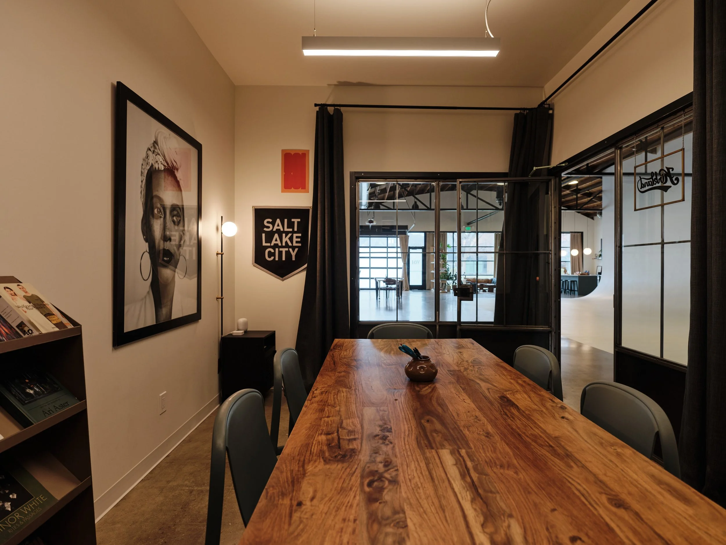 A cozy interior space with a large wooden table, surrounded by six chairs. The room features framed art on the wall, with a prominent black and white portrait of a woman with pink hair, a 