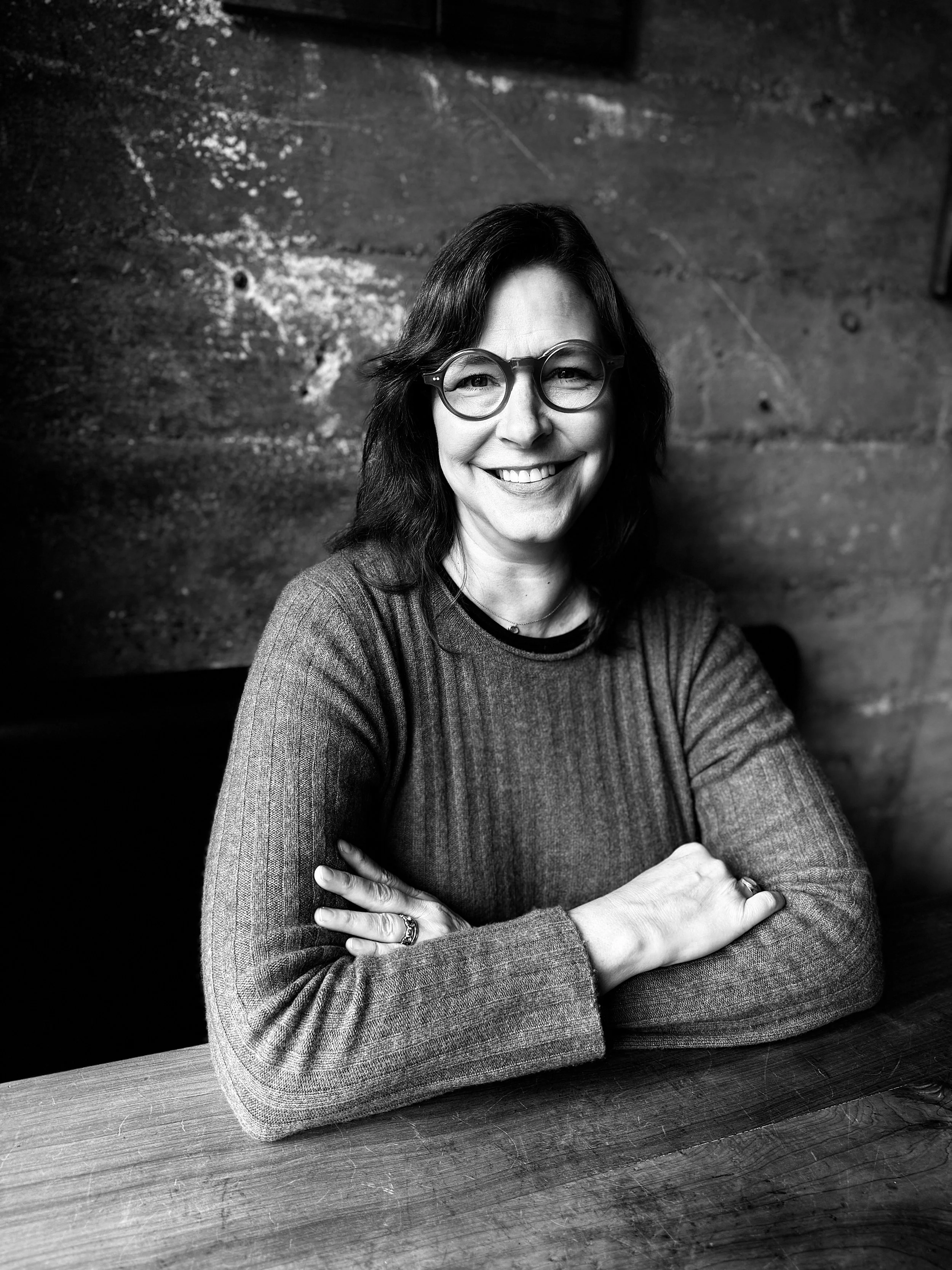 Black and white portrait of a smiling woman with glasses and dark hair, wearing a sweater, seated at a wooden table.