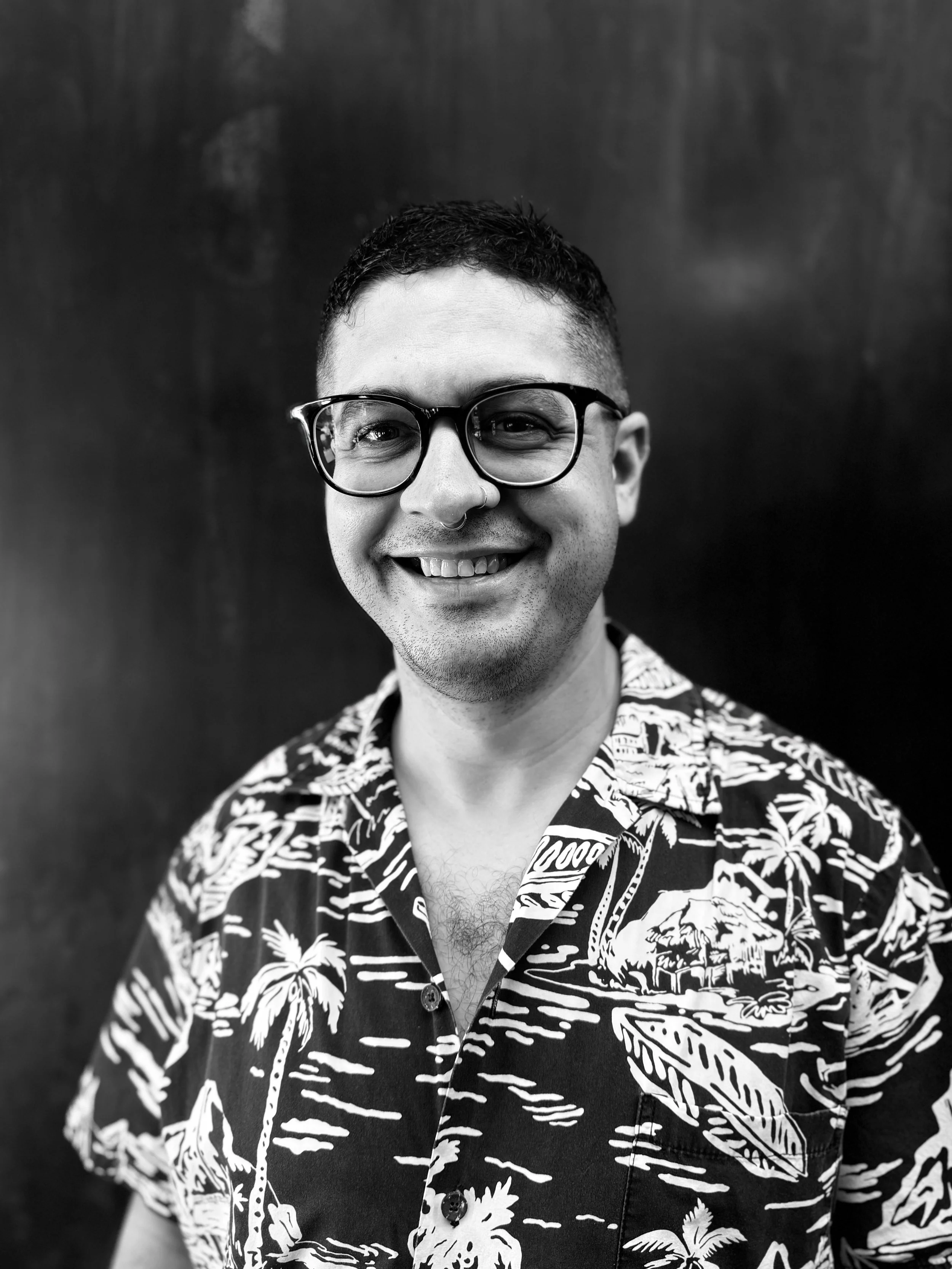 Black and white portrait of a smiling man with glasses, wearing a Hawaiian shirt with tropical patterns, against a dark background.