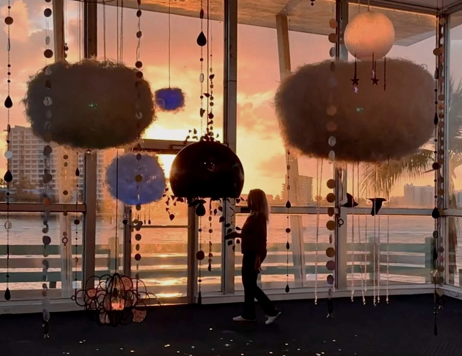 Silhouette of a person standing inside room with large windows, hanging sculptural lanterns objects, during sunset over Biscayne Bay and city skyline.