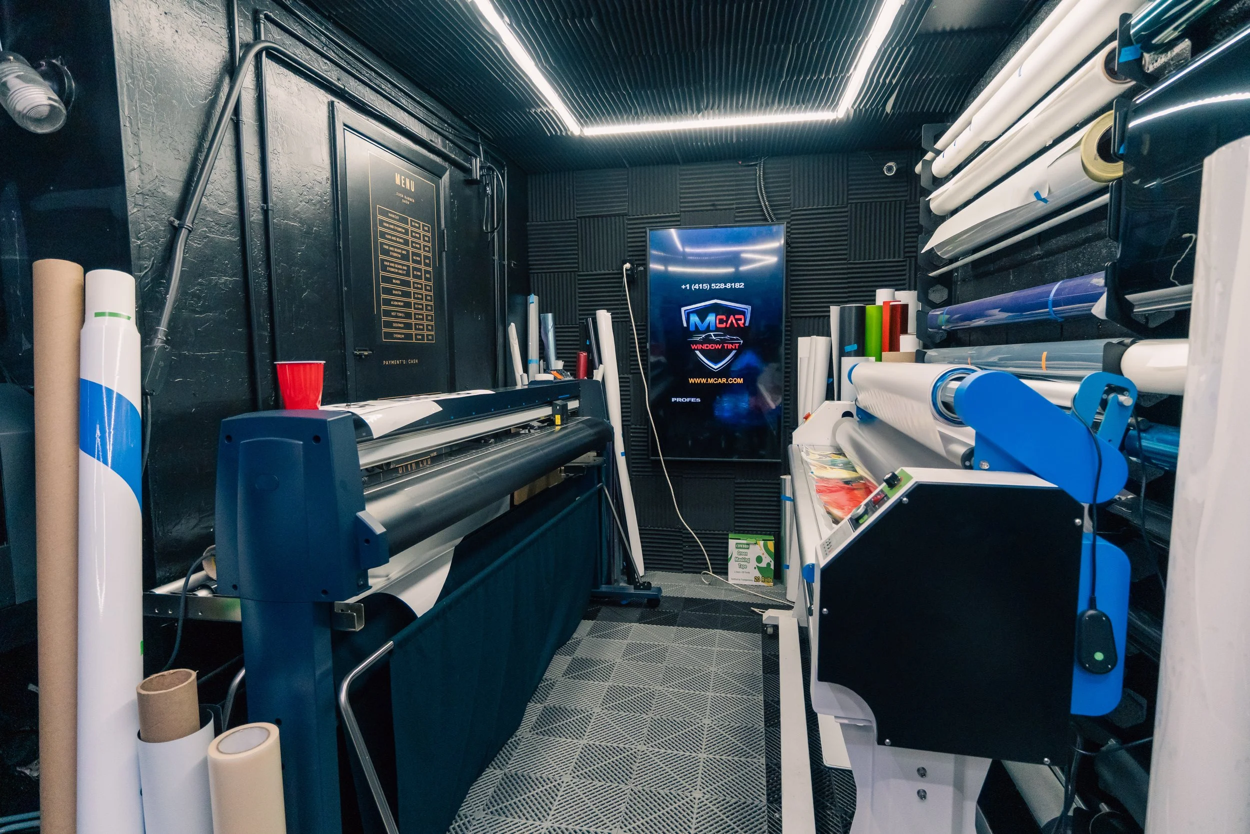 An interior of a print shop or workshop with rolls of vinyl wrapping material, a cutting machine, and a digital display screen on a black wall.