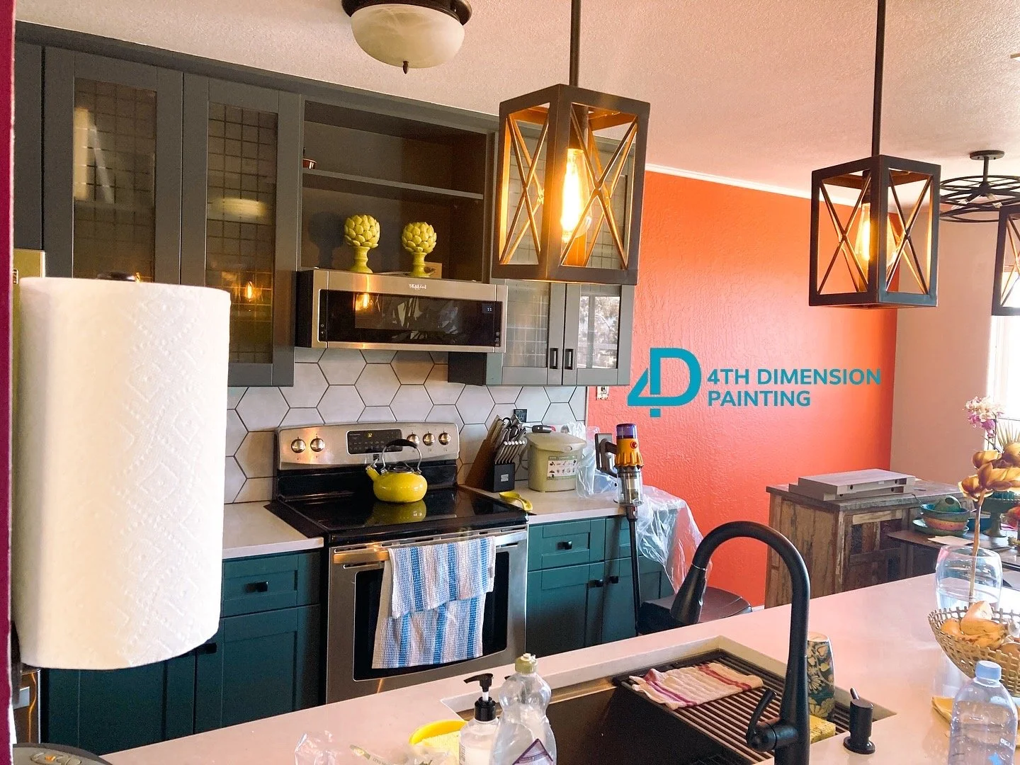 Kitchen with dark teal cabinets, stainless steel stove, microwave, and hexagonal backsplash tiles. Orange wall with wood furniture and modern black pendant lights.