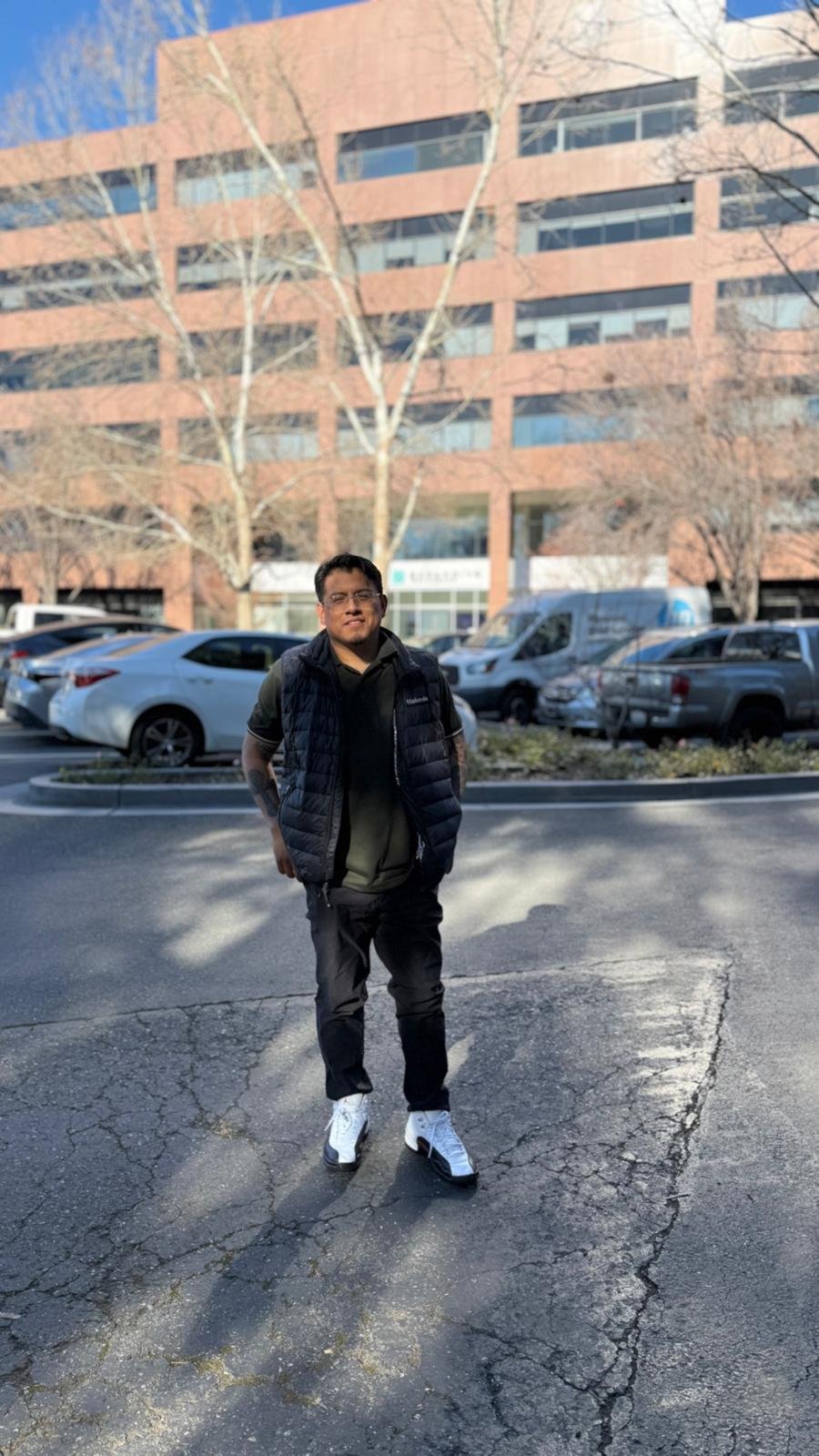 A man standing on a cracked asphalt parking lot in front of a row of parked cars, with a large office building and leafless trees in the background, during what appears to be winter or early spring.