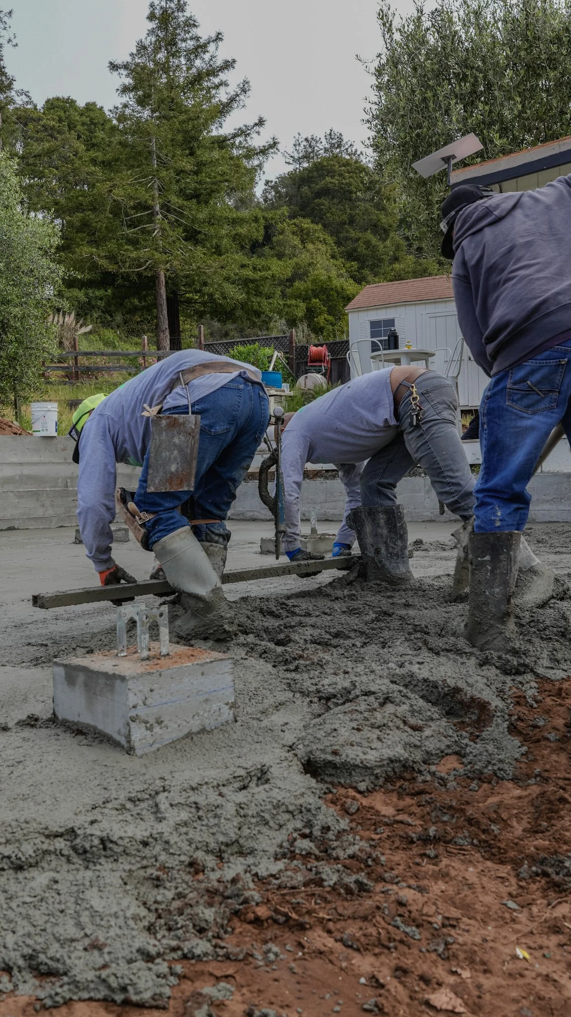 Three workers pouring and leveling concrete on a construction site outdoors, with trees and a shed in the background.