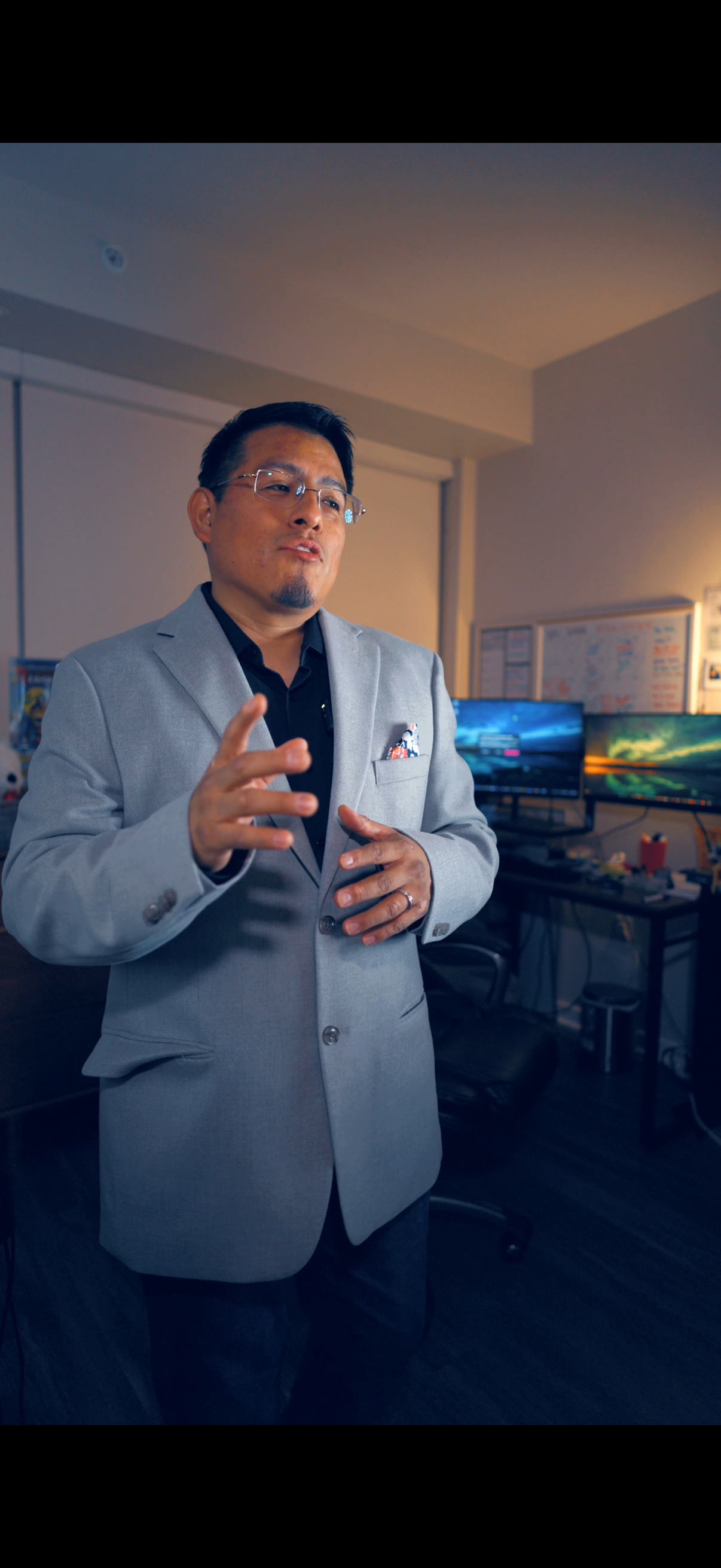 A man in a light gray suit and black shirt standing in an office, gesturing with his right hand, with multiple computer monitors in the background.