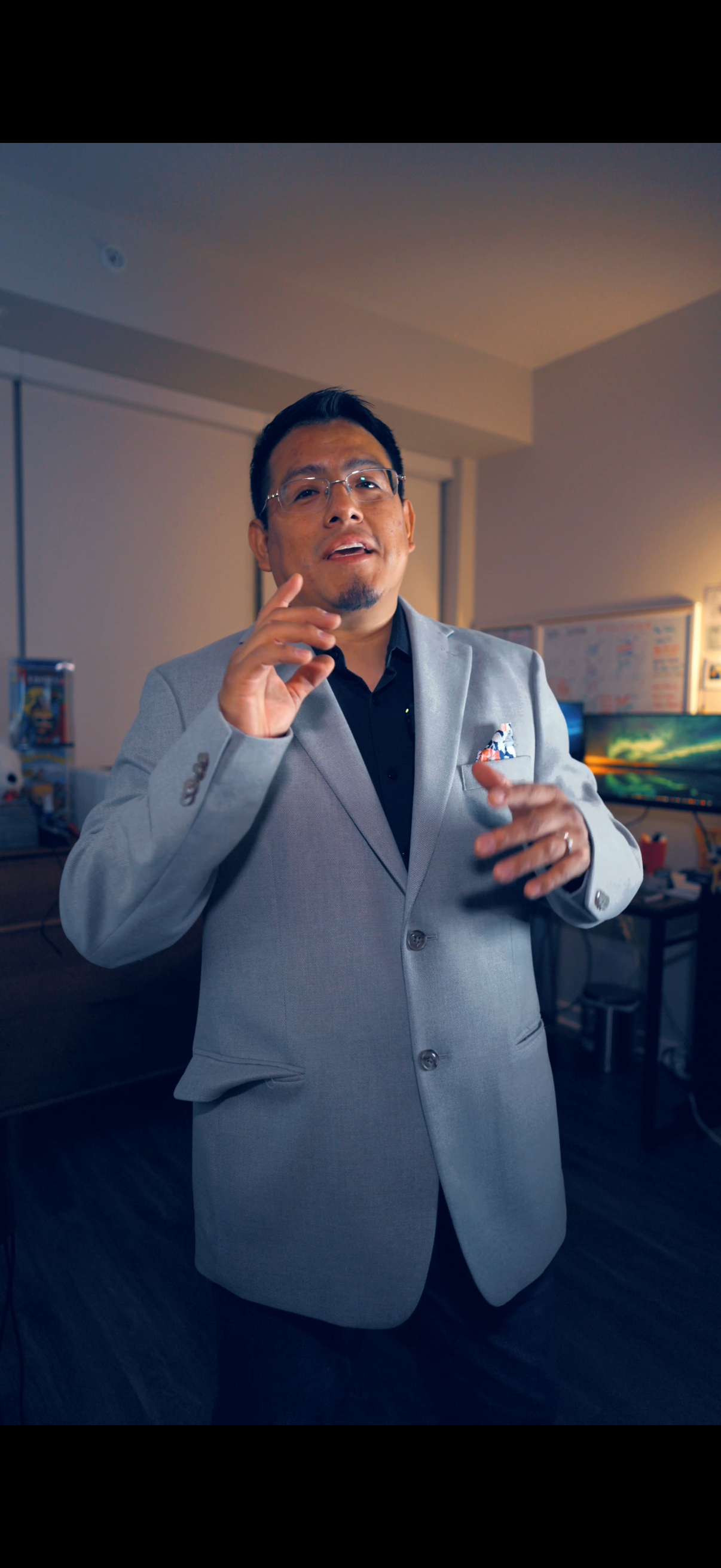 A man wearing glasses and a light gray suit jacket, standing in a room with computer monitors and whiteboards in the background.