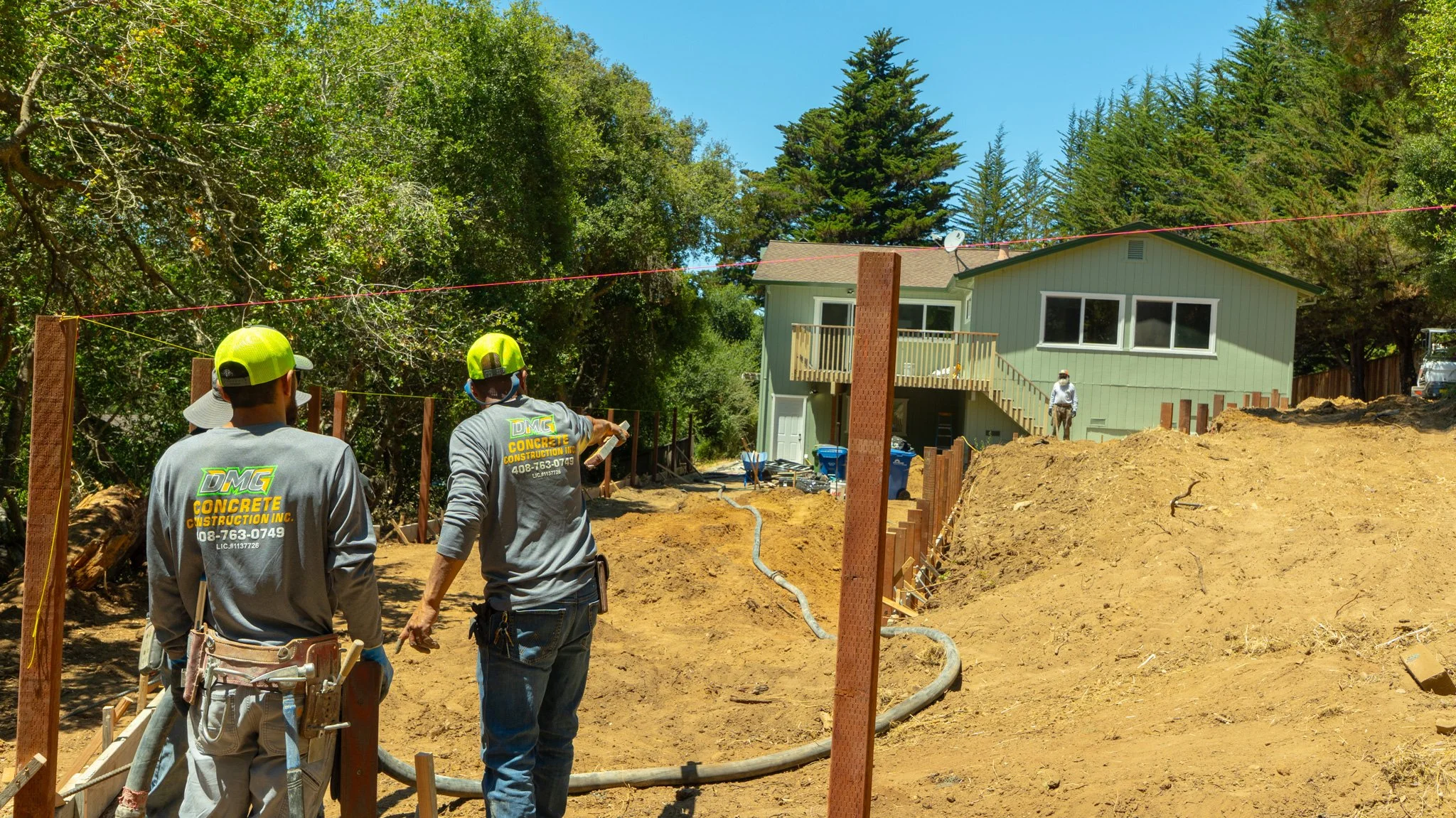 Construction workers working on a building site in front of a green house, with trees in the background, dirt, construction equipment, and a partially built fence.