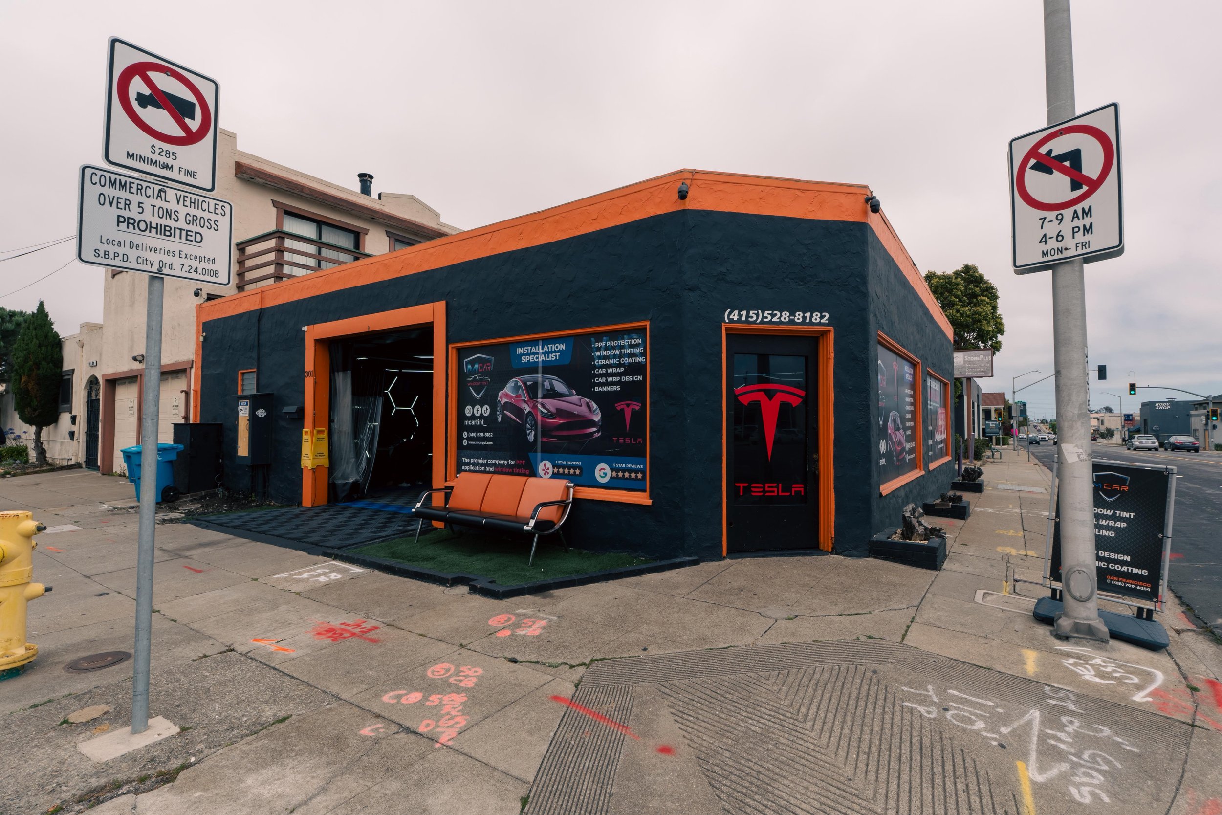A dark gray and orange storefront with Tesla branding on the windows and door. There are signs on the building advertising installation services for window tint and paint protection. A bench is in front of the store, and there are street signs indicating parking restrictions nearby. The sidewalk has various numbers and markings painted on it.