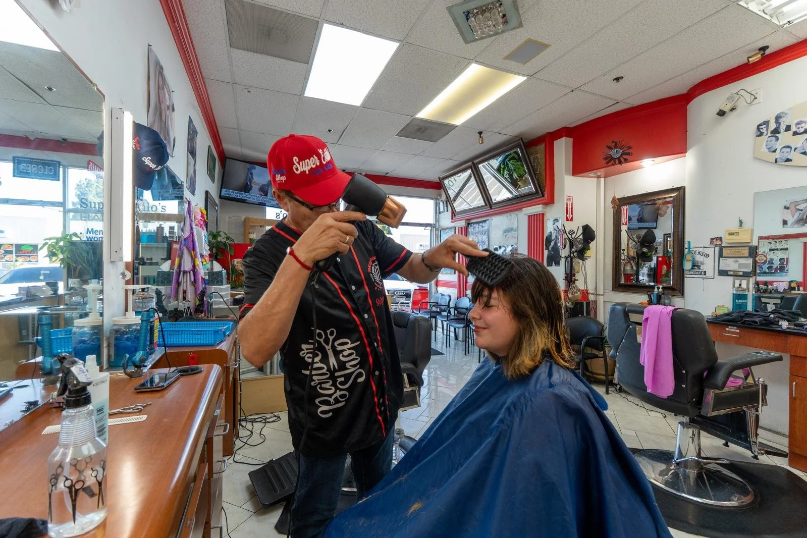 Barber styling a woman's hair in a salon with various mirrors, chairs, and hair styling tools visible.