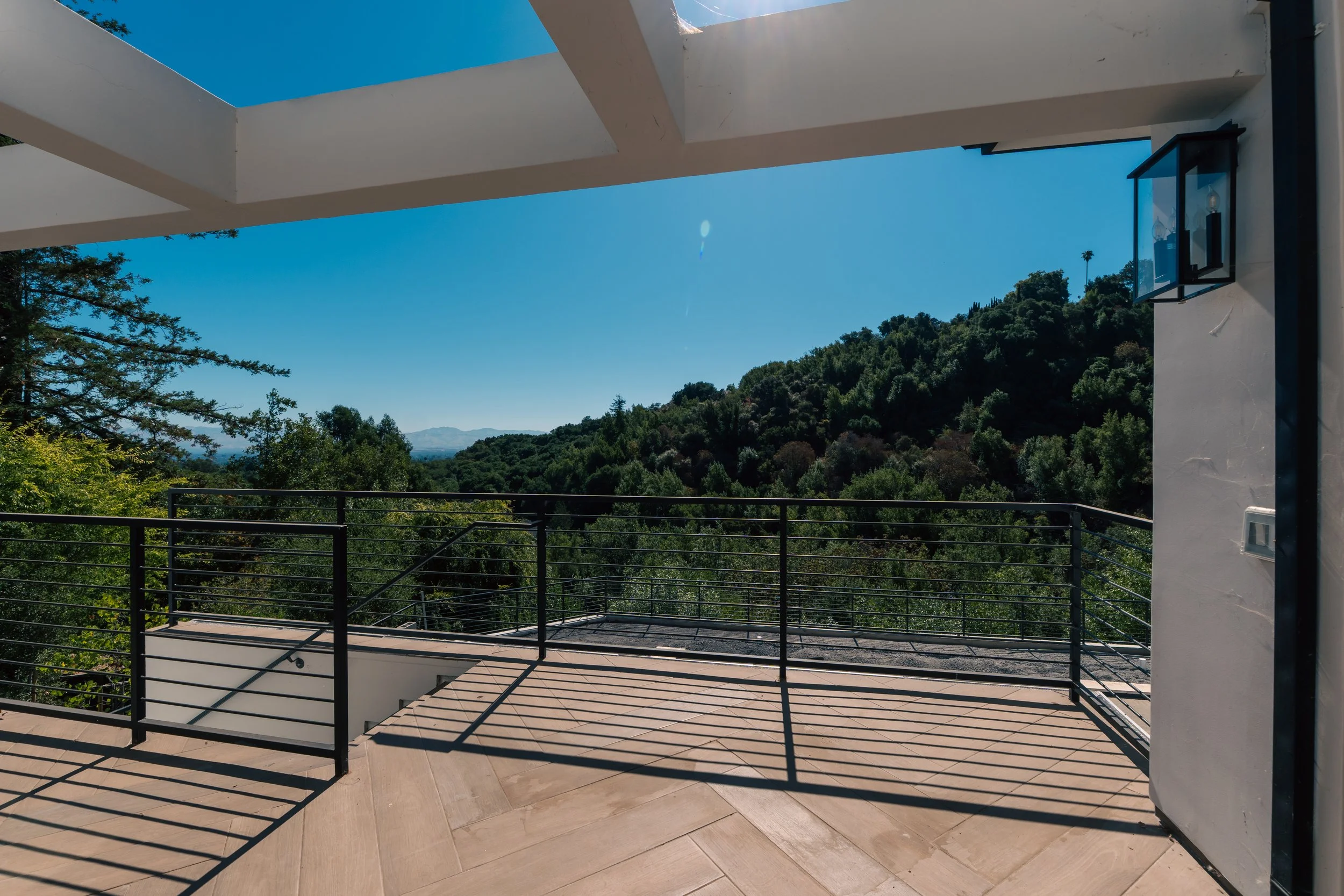 View of a balcony with black metal railing overlooking green forested hills and distant mountains under a clear blue sky.