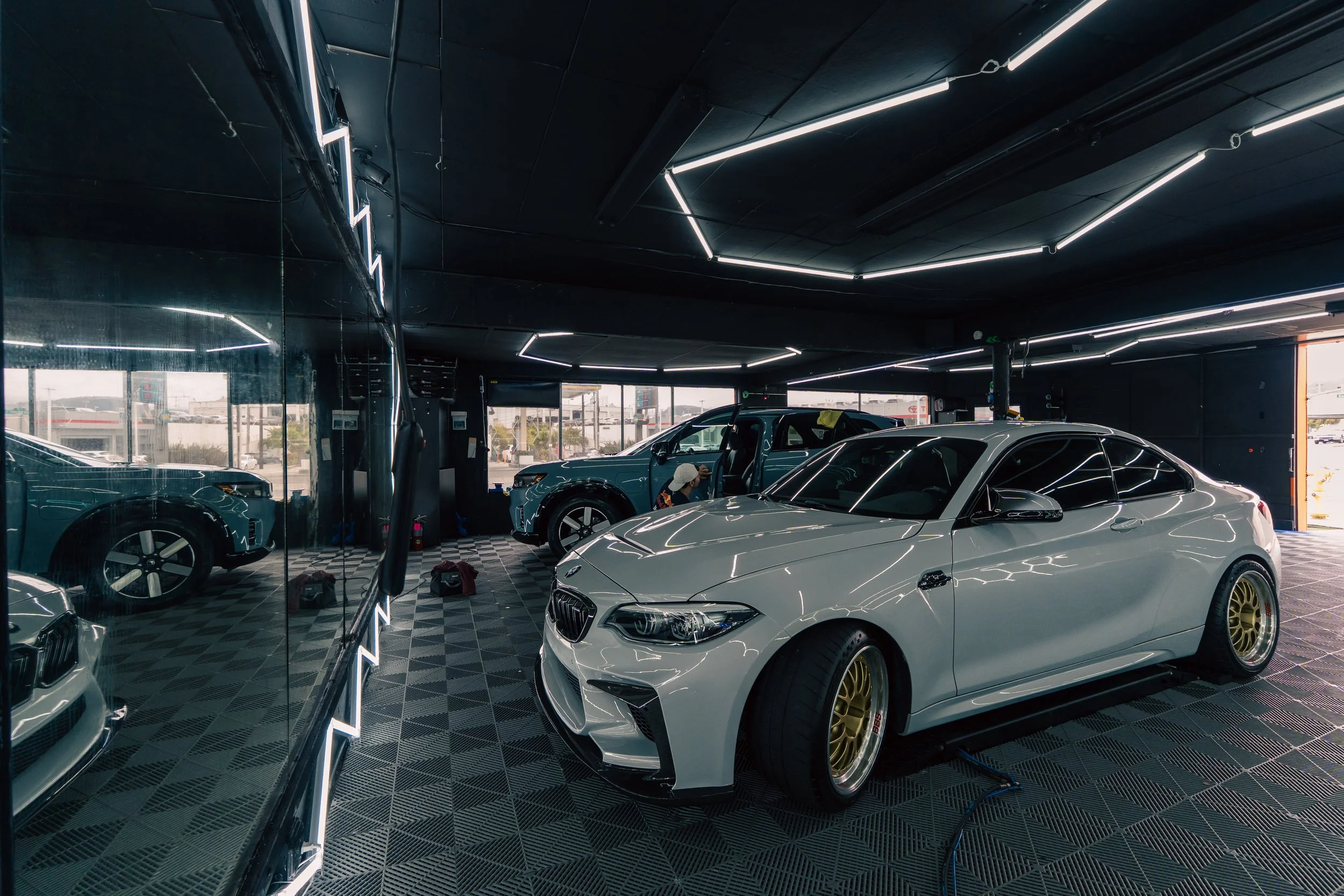 A showroom with luxury cars, including a white sports car with gold rims in the foreground, and a silver SUV in the background, reflected in the large glass window.