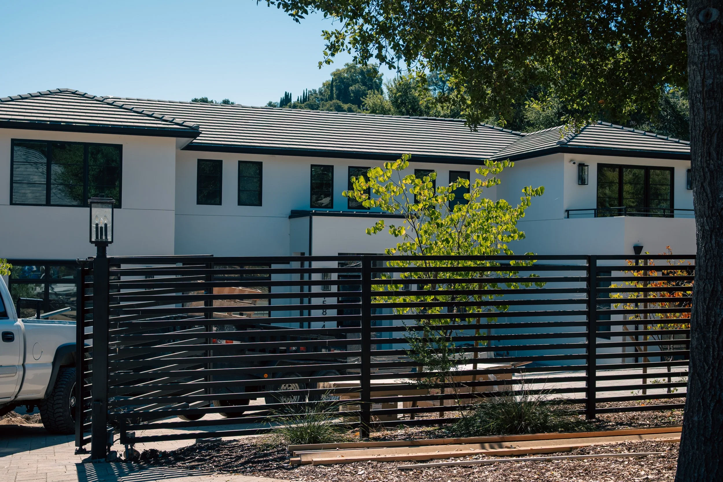 Modern two-story white house with black-framed windows, surrounded by a black fence, trees, and plants, under a clear blue sky.