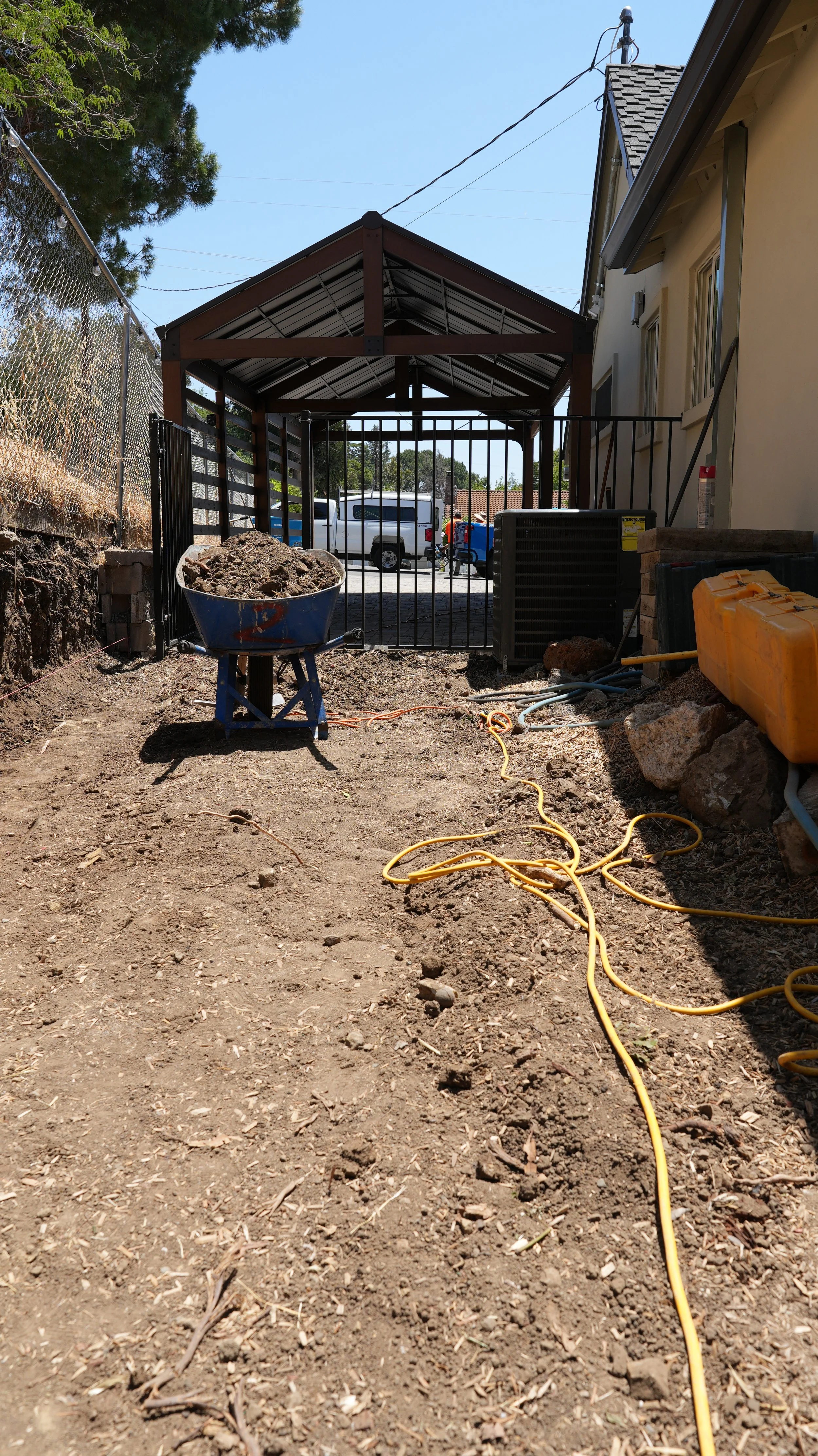 A backyard construction site with dirt ground, a wheelbarrow filled with dirt, yellow extension cords, and construction materials near a house. A decorative metal gate and a covered patio are visible in the background, along with parked vehicles.