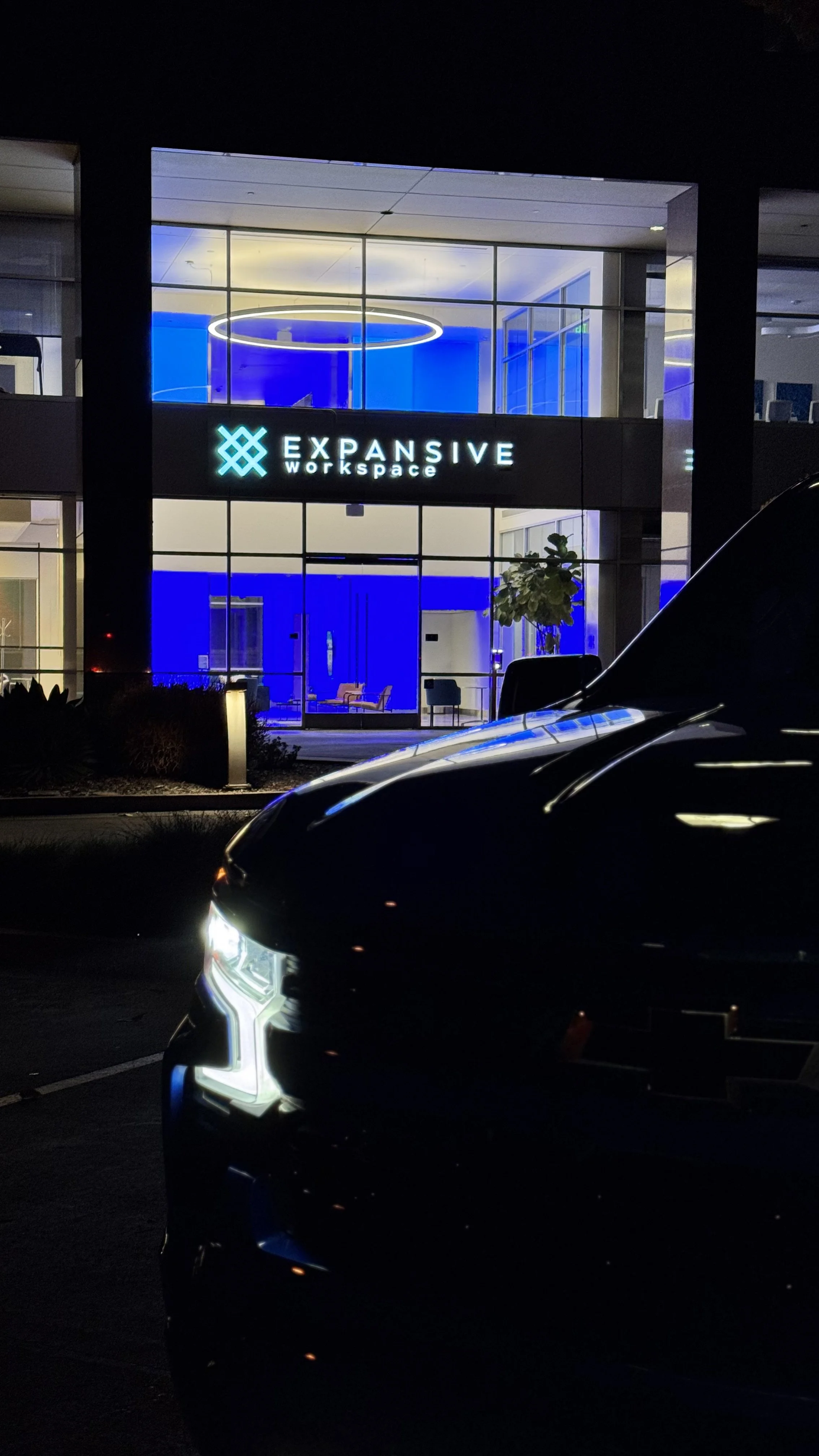 Night view of a modern two-story office building with a sign that reads 'Expansive Workspace.' The building has large glass windows illuminated with blue lights, and a black car is parked in the foreground.