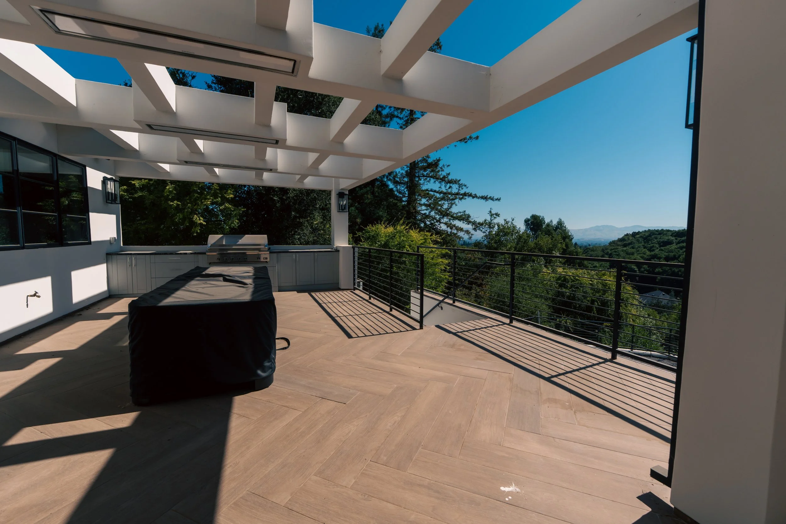 Modern outdoor balcony with wooden flooring, black metal railing, and a built-in grill, overlooking a lush green landscape and blue sky.
