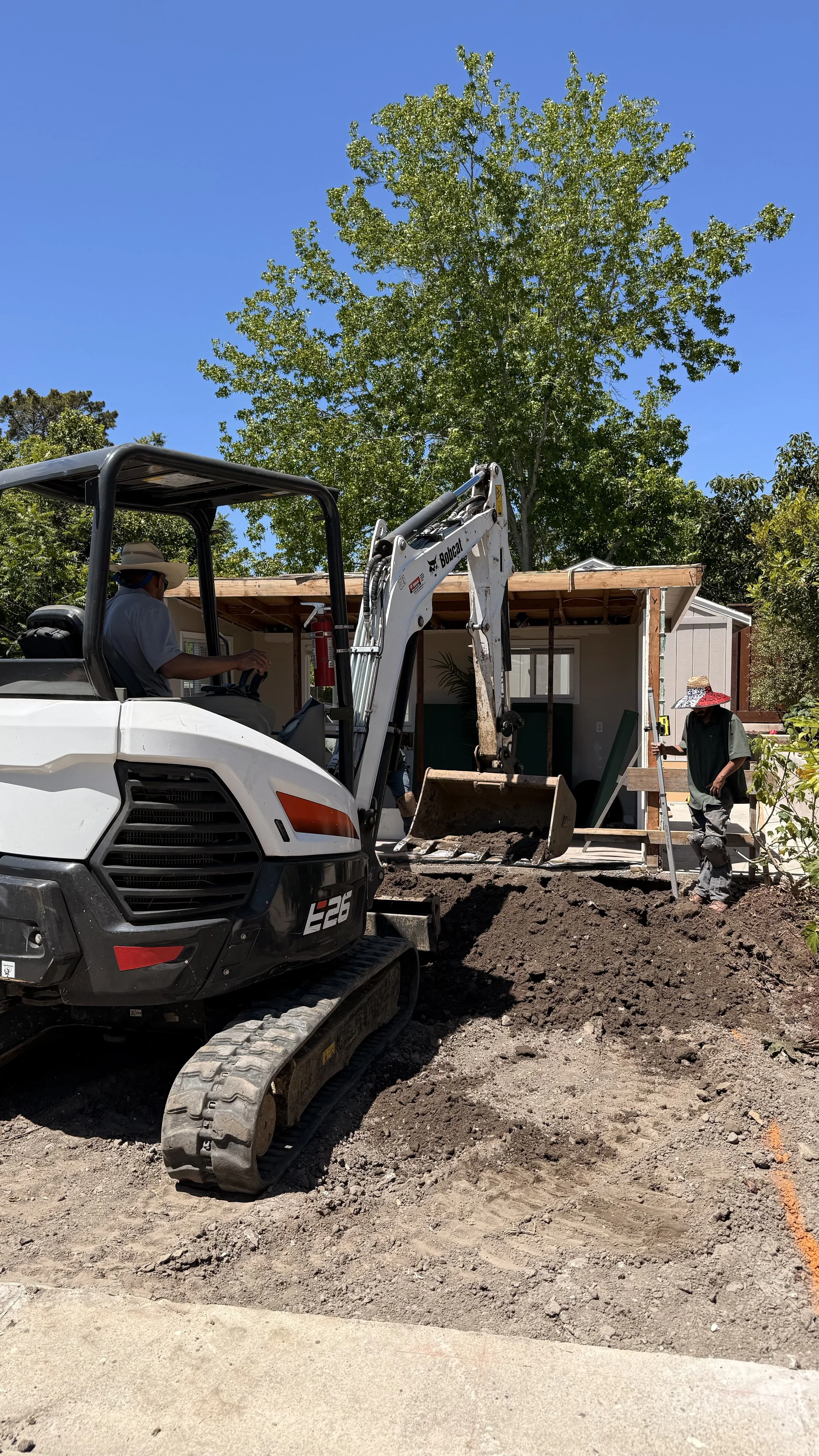 Construction workers operating a Bobcat mini excavator and using a level at a home renovation site with dirt and a partially built wall.