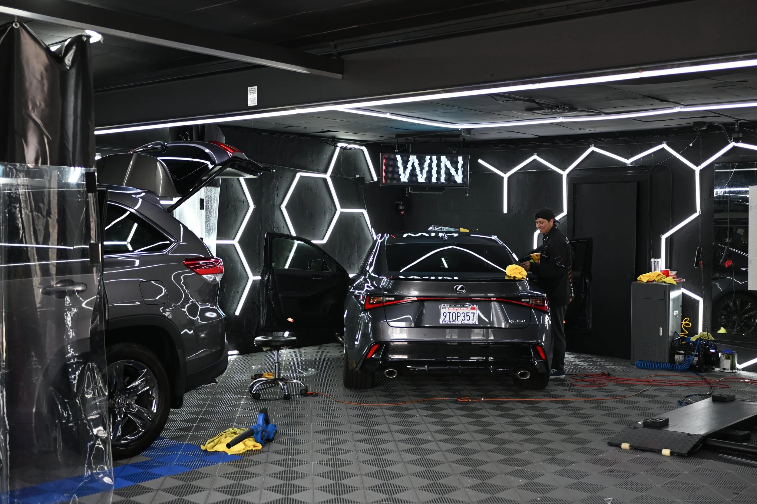 A mechanic cleaning a black Lexus in a modern, black-themed car detailing garage with hexagonal light patterns and a digital sign that reads 'WIN'.