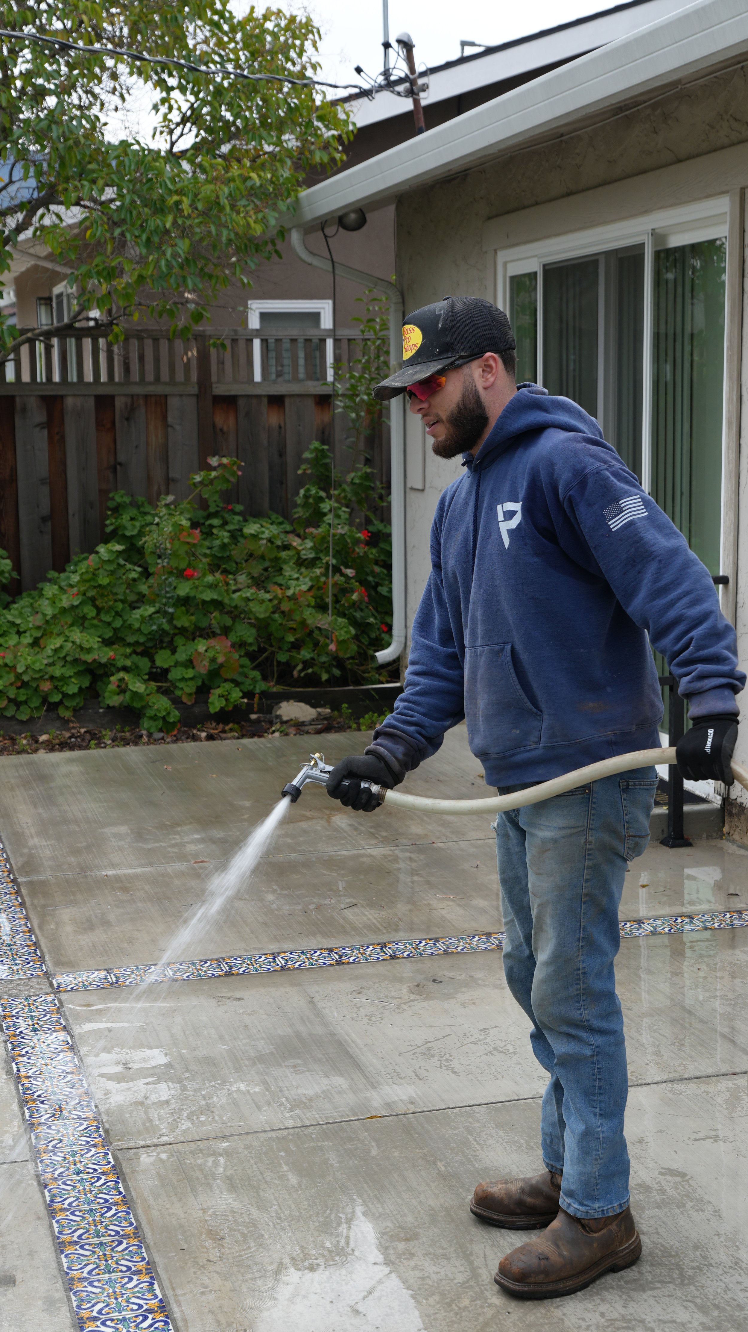 Man wearing a black cap, sunglasses, blue hoodie, and jeans power washing a concrete patio outside a house.
