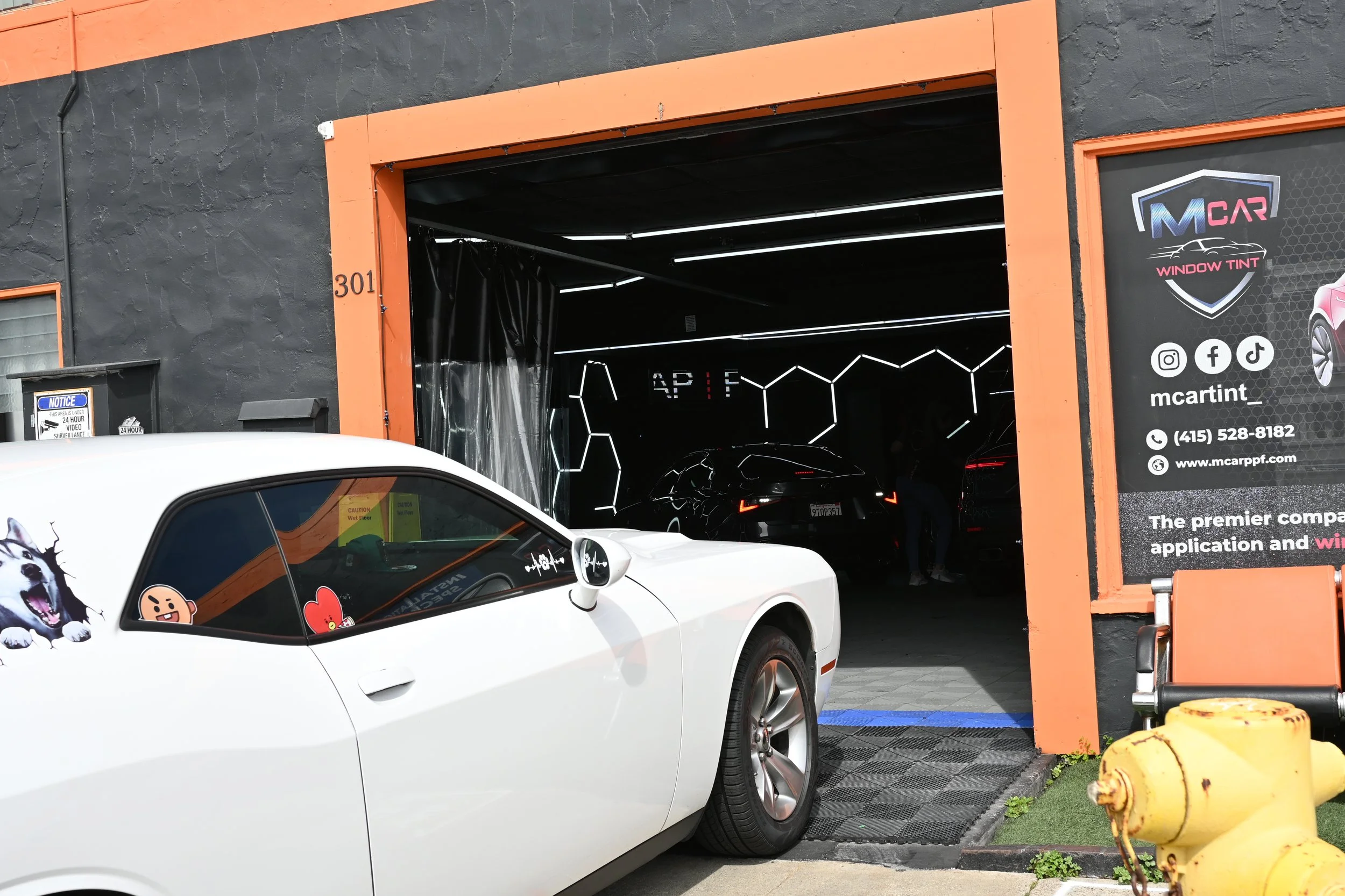 A white sports car with cartoon stickers on the back window parked outside an auto detail shop with an open garage door. Inside the garage, a black car is being serviced or cleaned, with neon decorative lighting on the walls.
