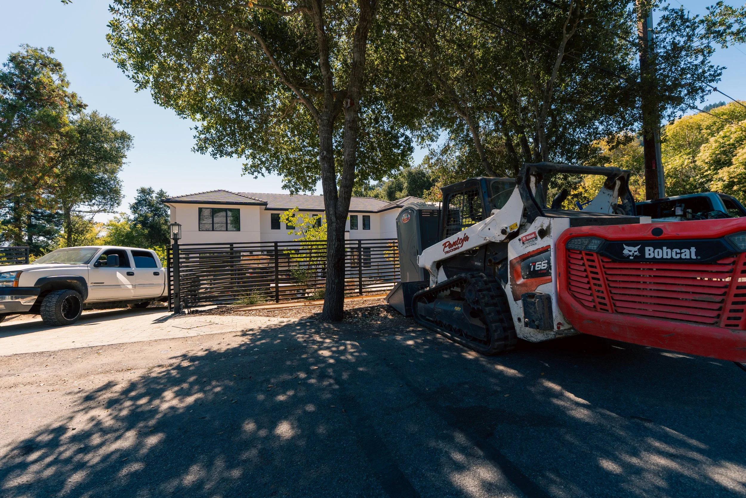 A modern two-story house with white exterior walls and black window frames set behind a black metal fence. In the foreground, there's a large tree casting shadows on the street, a white pickup truck, and a red and white Bobcat T66 tracked utility veh