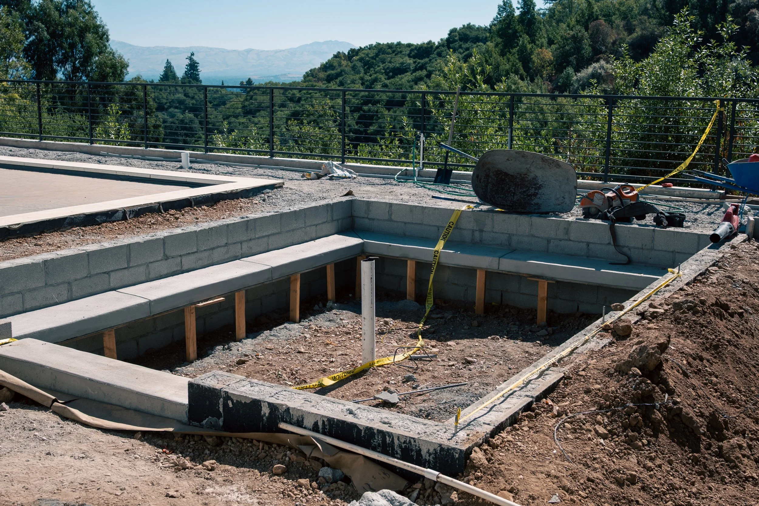 Construction site with a concrete pool surrounded by a wooden frame, caution tape, construction tools, and equipment, with a view of trees and mountains in the background.